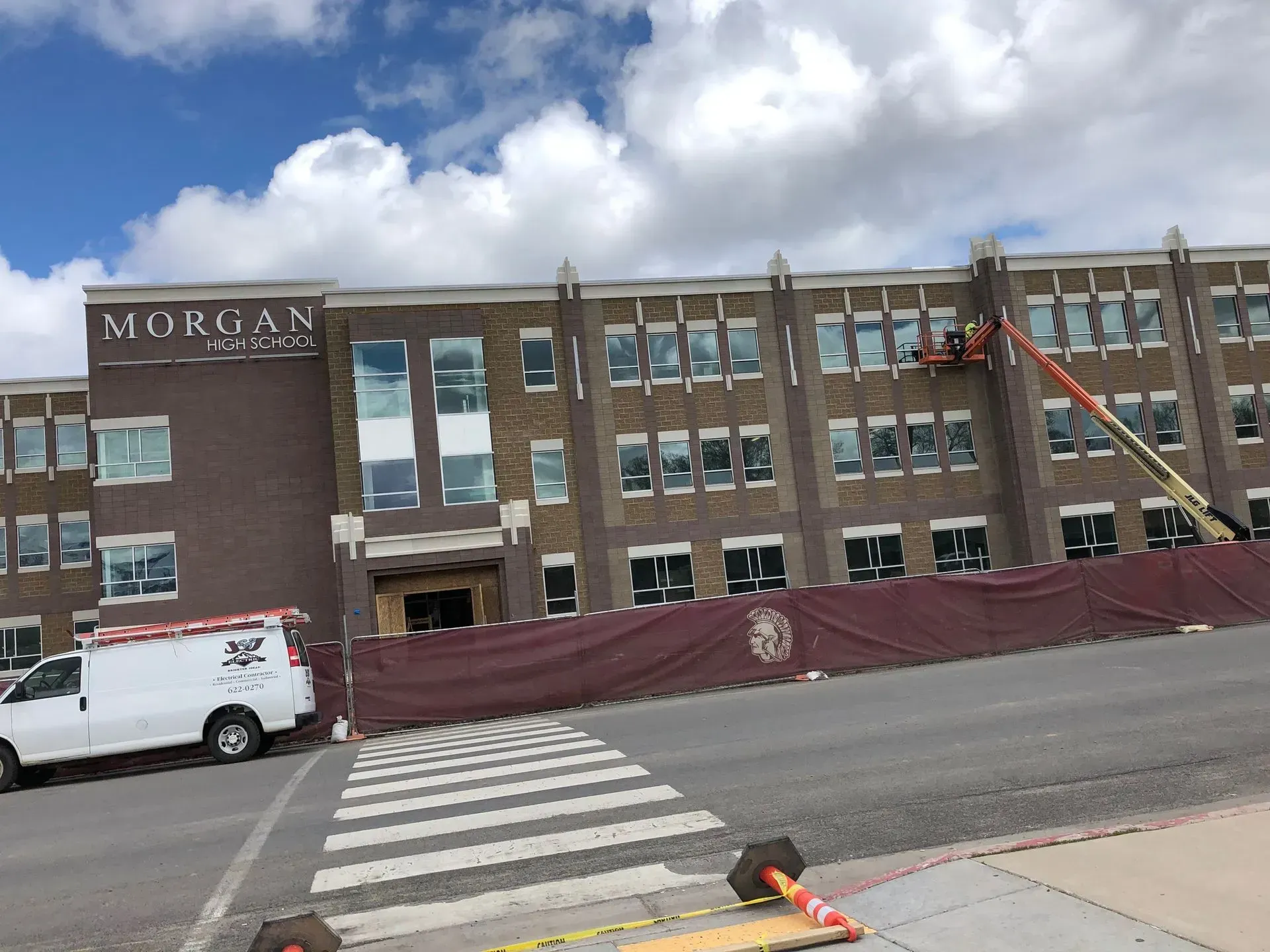 Construction workers using a lift at the brick Morgan School building, fronted by a construction fence and a white van.