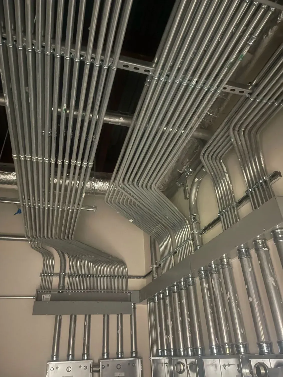 A complex array of numerous parallel silver electrical conduits running along a ceiling into metal junction boxes.