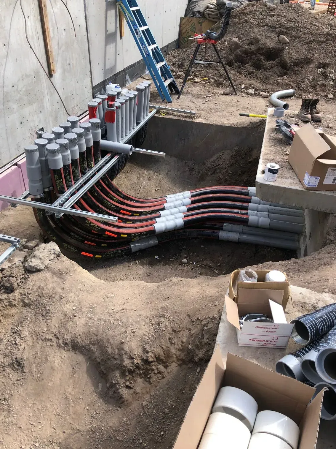 Multiple black pipes curve from a concrete foundation into an excavated dirt trench at a construction site.