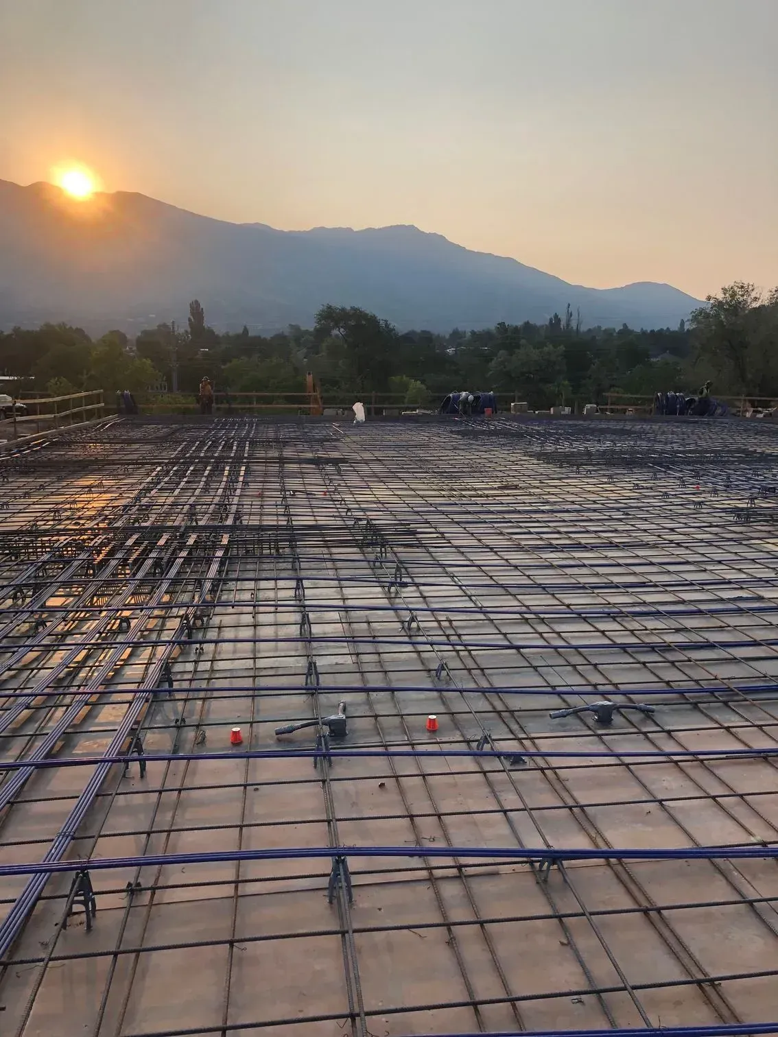 Construction site showing a rebar grid and blue tubing laid on concrete before a pour at sunset with mountains behind.