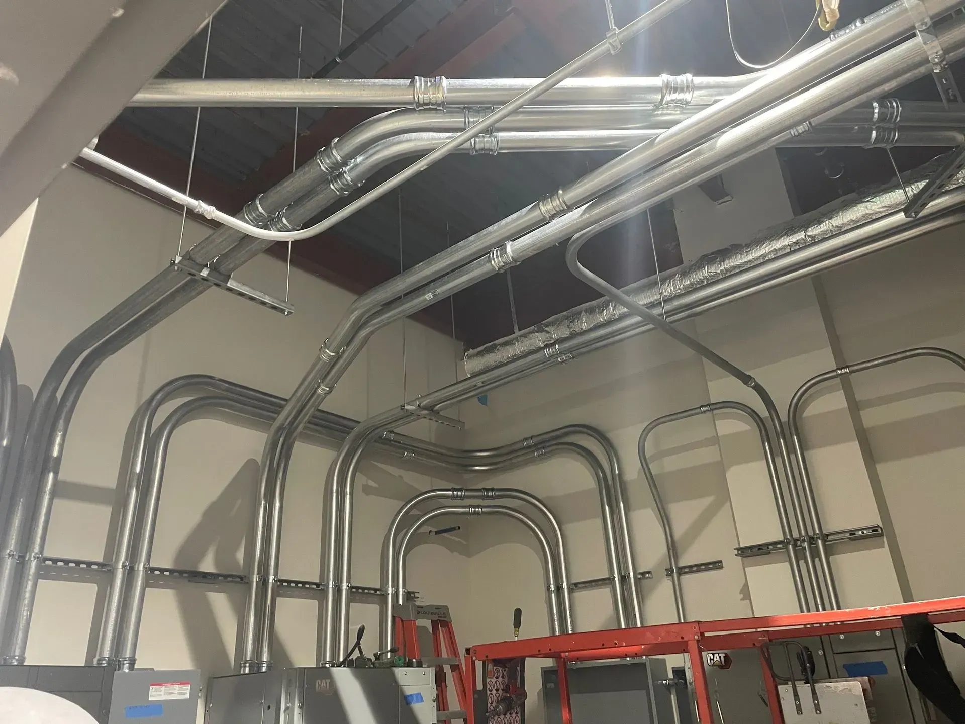 Numerous metal electrical conduits running along a wall and ceiling in an industrial setting.