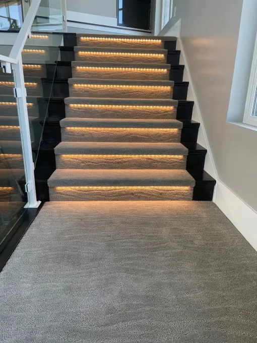 A staircase with grey carpeted steps and integrated warm lighting under each tread, flanked by a glass and white railing.