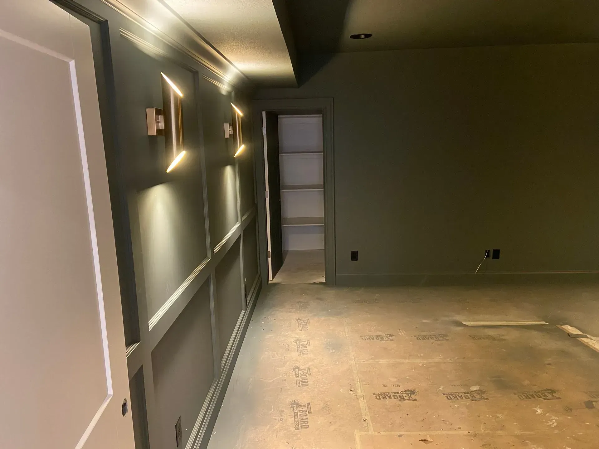 A room with gray wainscoted walls, two wall-mounted light fixtures, an open doorway to a shelf-lined closet, and subfloor.