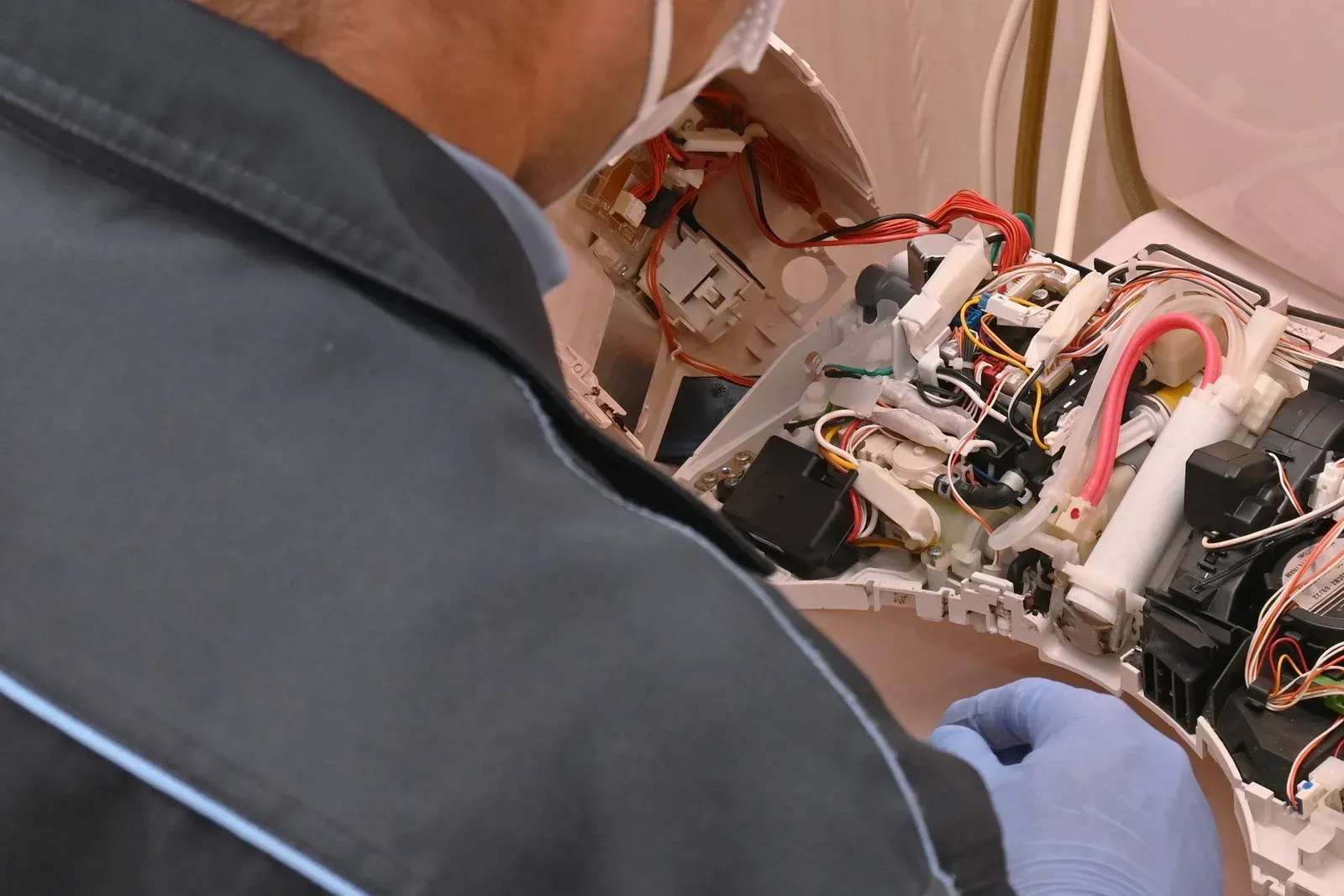 A technician in a blue uniform and gloves repairs the exposed internal wiring and components of a smart toilet seat.