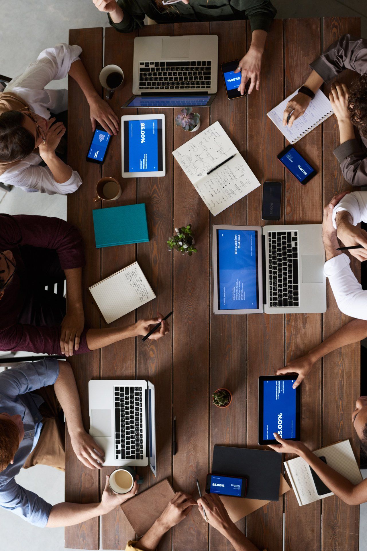 A group of people are sitting around a wooden table with laptops and tablets.