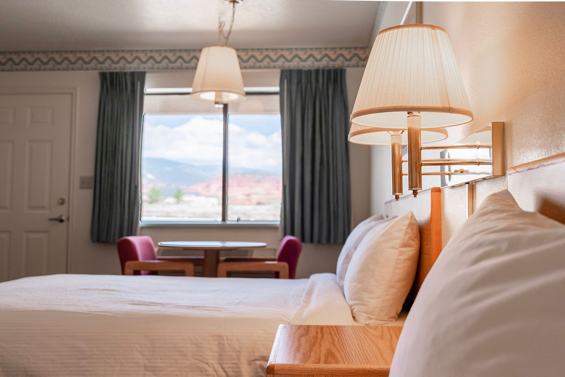 A hotel room with a large bed , two lamps , a table and chairs.