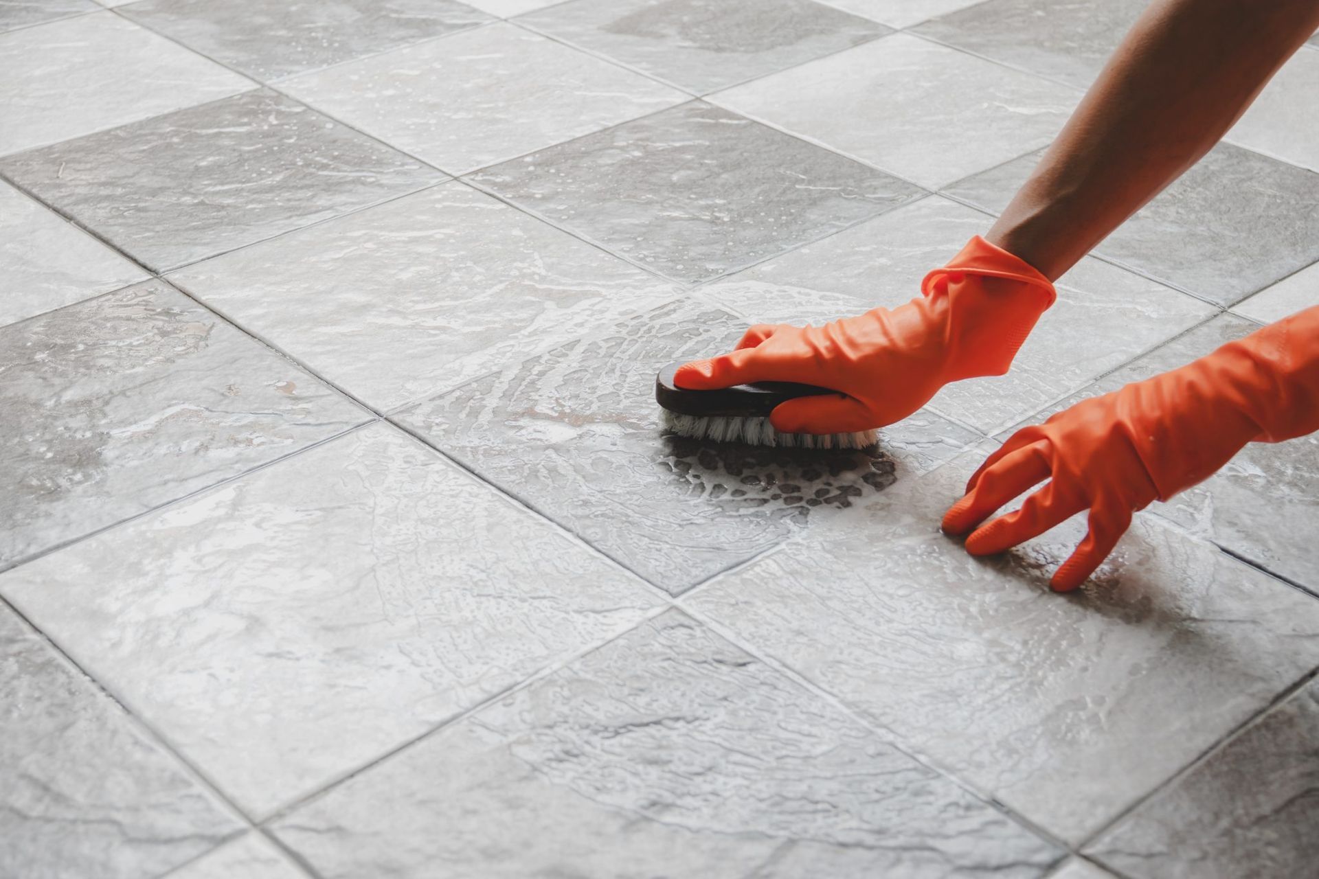 Hands in orange gloves scrubbing a gray tiled floor with a brush