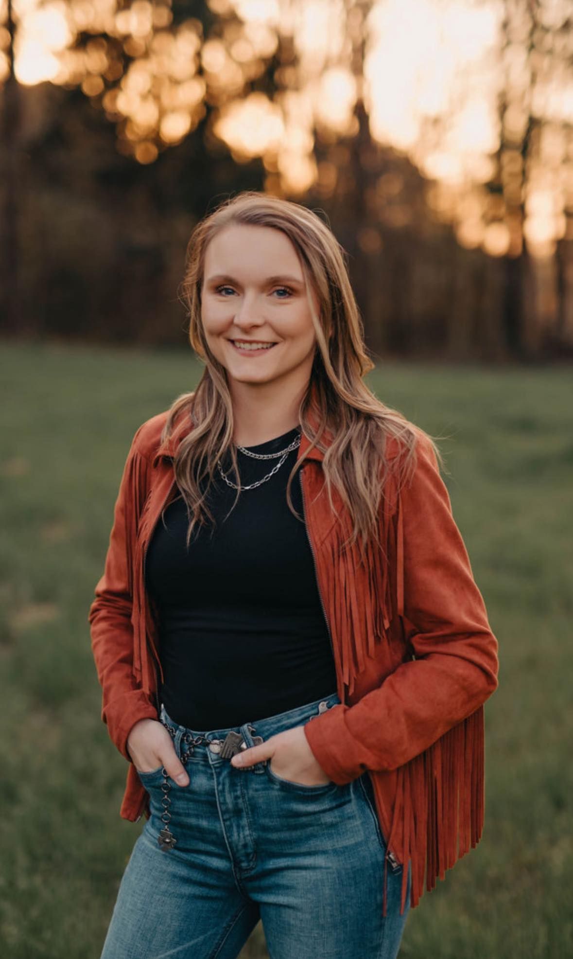 Woman in a rust-colored fringe jacket and jeans smiles, hands in pockets, set against a field at sunset.