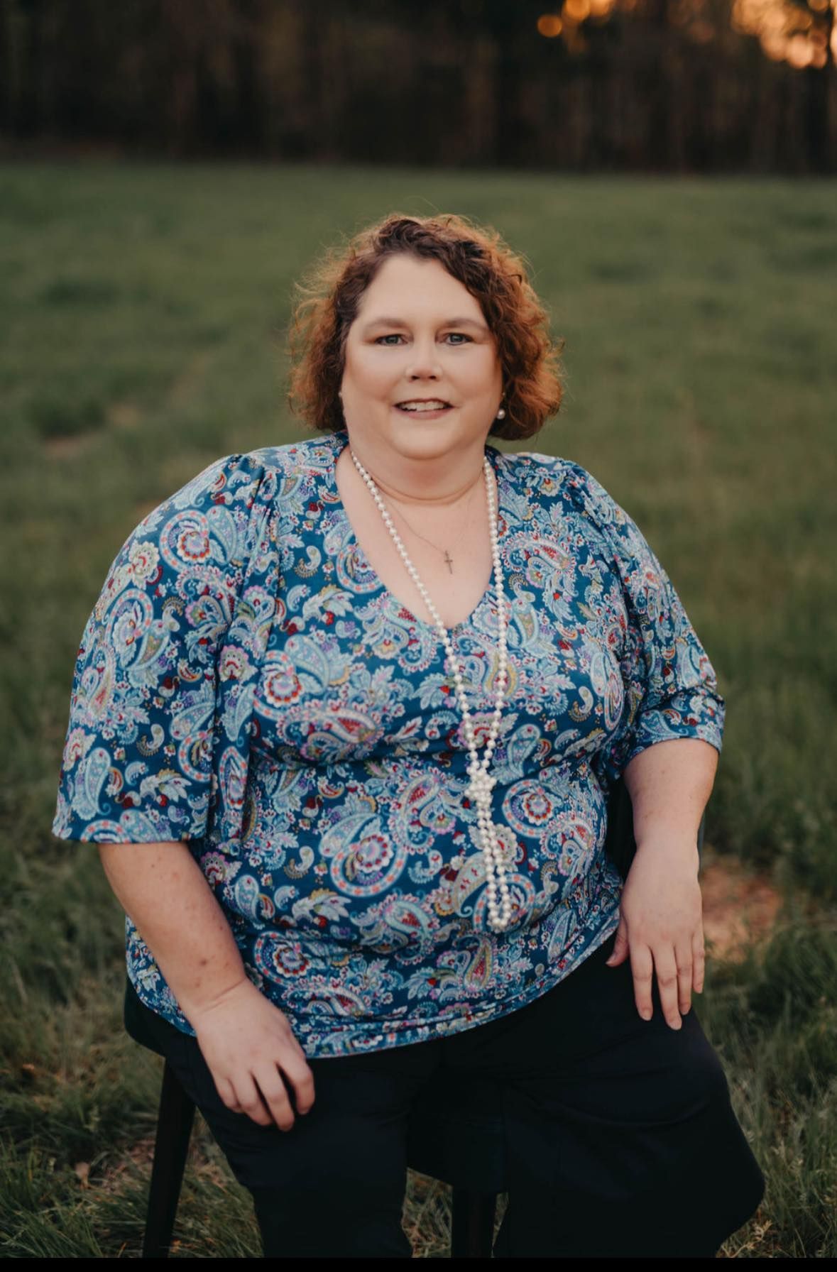 Woman with curly brown hair in blue paisley top and pearl necklace, smiling, seated outdoors.