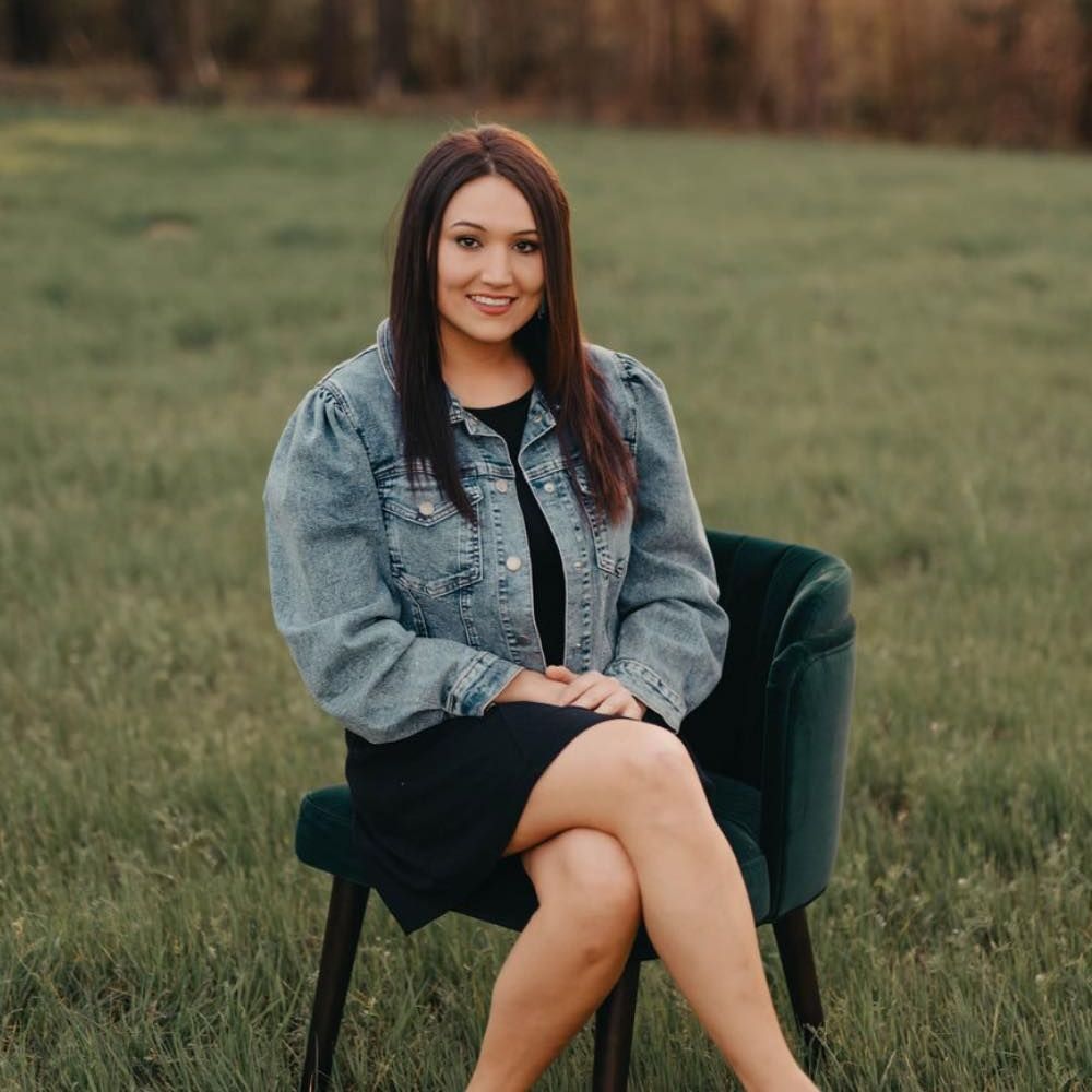 Woman with dark hair, wearing a denim jacket and black dress, sits on a teal chair in a field, smiling at the camera.