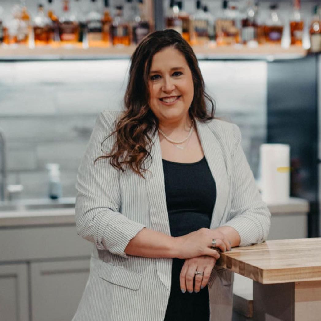 Woman smiling, leaning on a counter in a bar. She wears a black top, pinstripe blazer, and jewelry.
