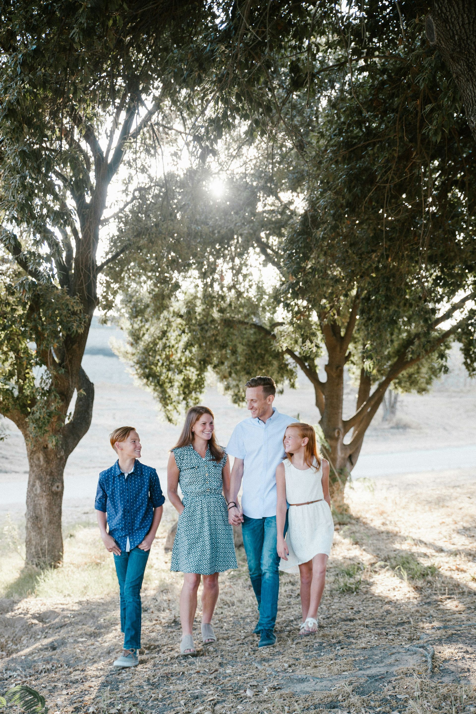 Family of four walking together under trees on a sunny day. They smile at each other.