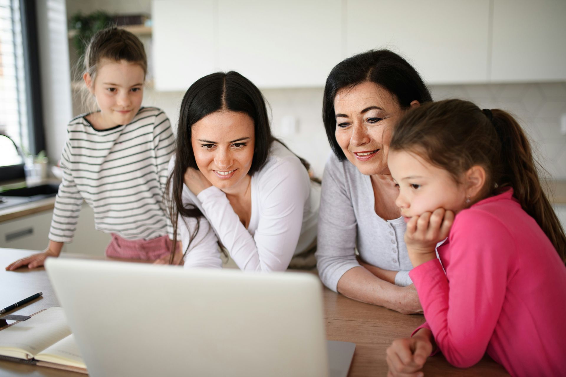 Four females, including two young girls, looking at a laptop screen in a bright kitchen; smiles and focused expressions.