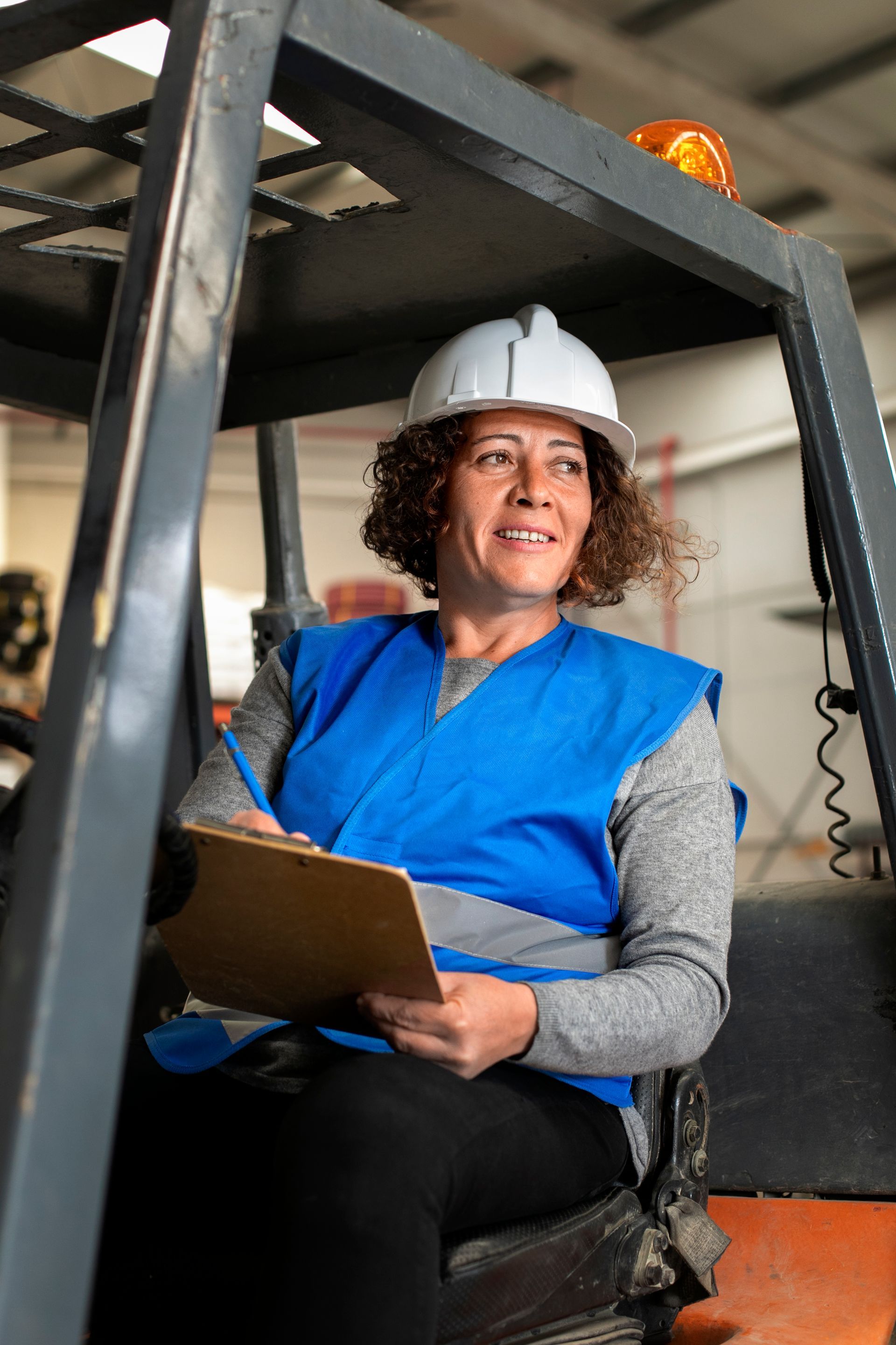 Woman in safety vest and hard hat sits in a forklift, holding a clipboard and smiling. Inside a warehouse.
