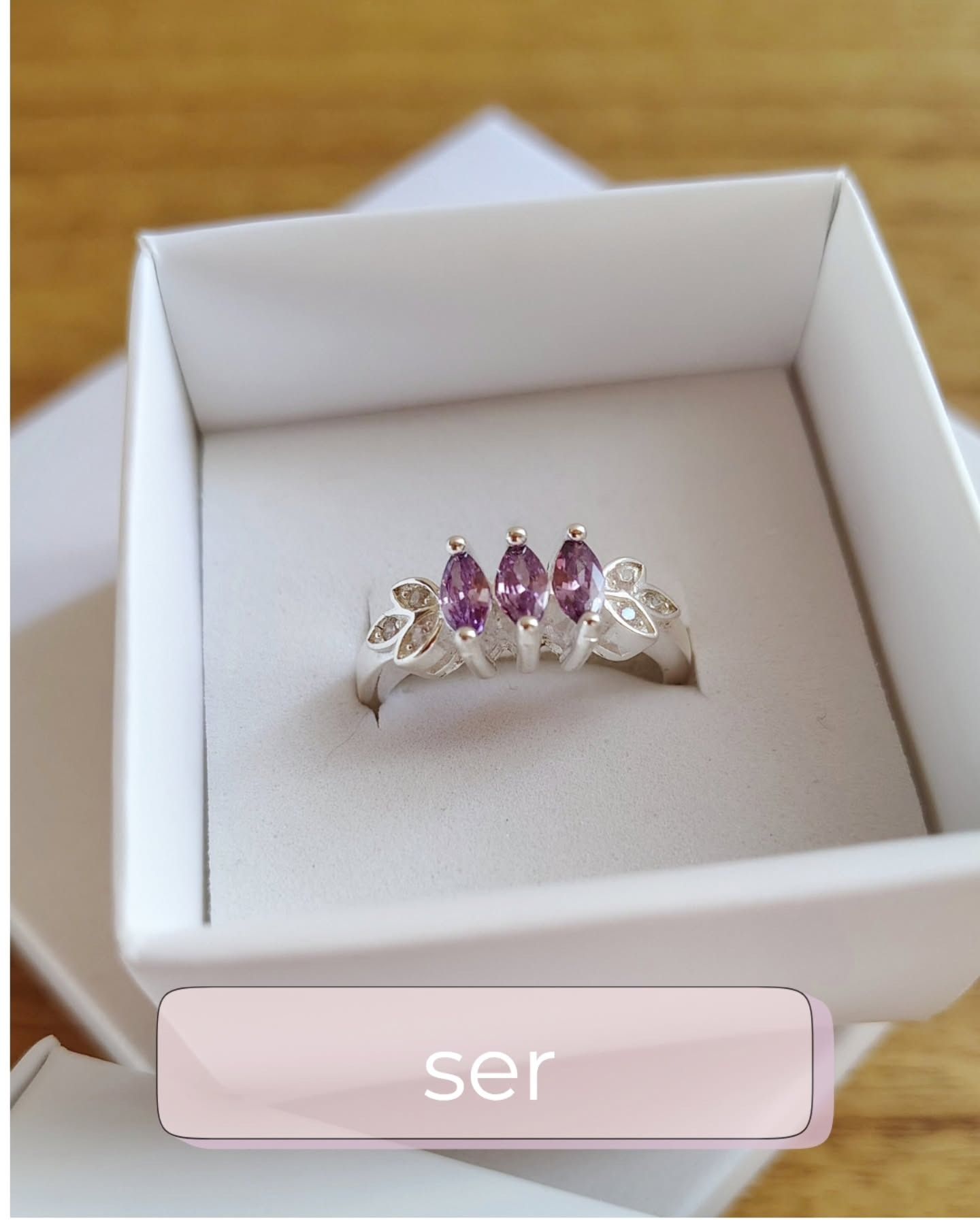 Anillo de plata con tres piedras de talla marquesa morada y detalles de hojas, en caja blanca.