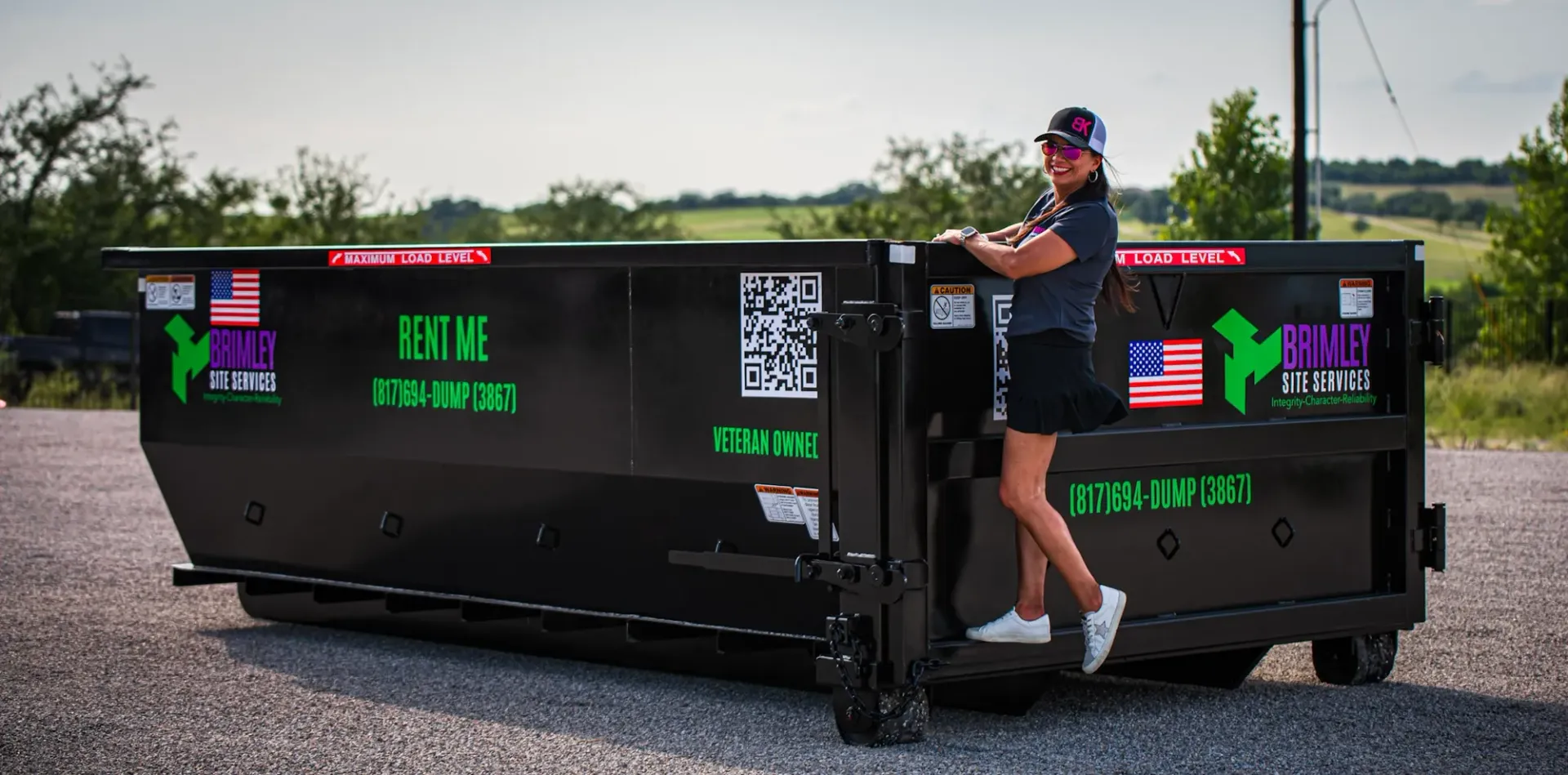 A woman is standing next to a dumpster on a gravel road.