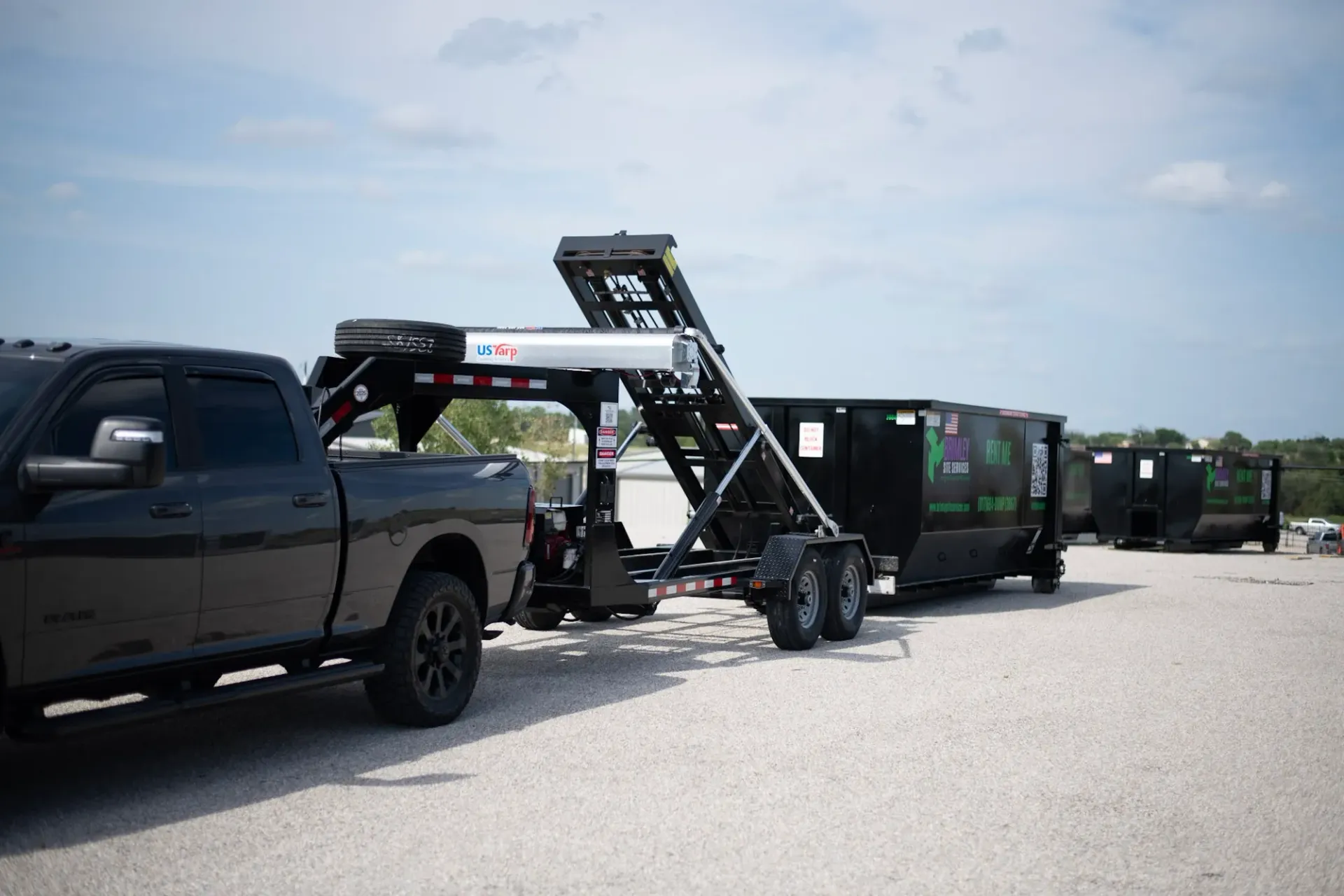 A black truck is towing a dump trailer in a gravel lot.
