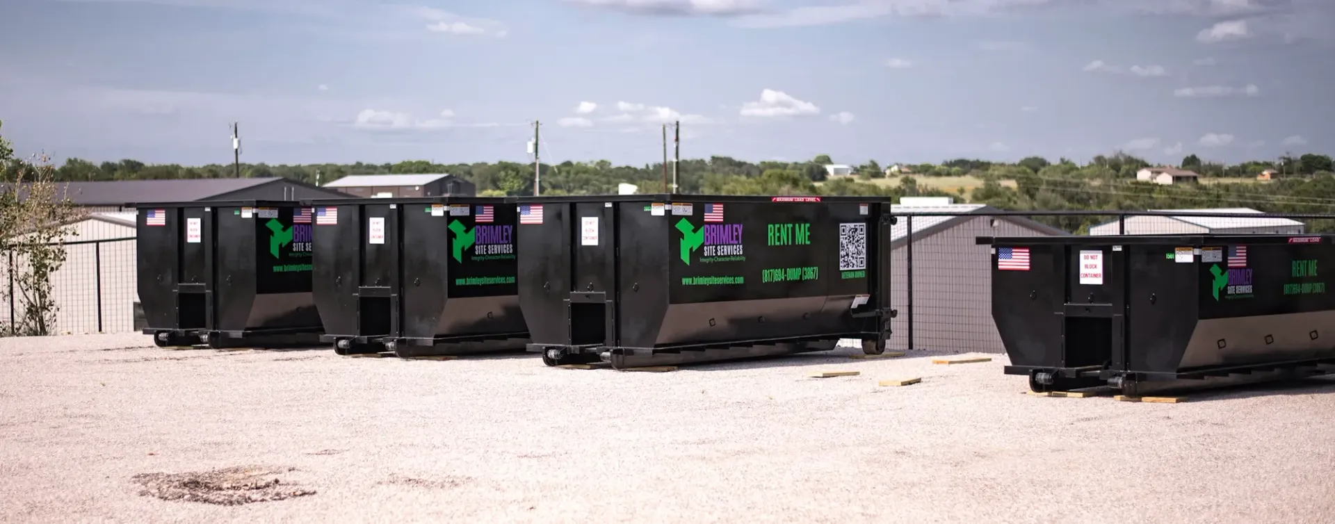 A row of dumpsters are parked in a gravel lot.