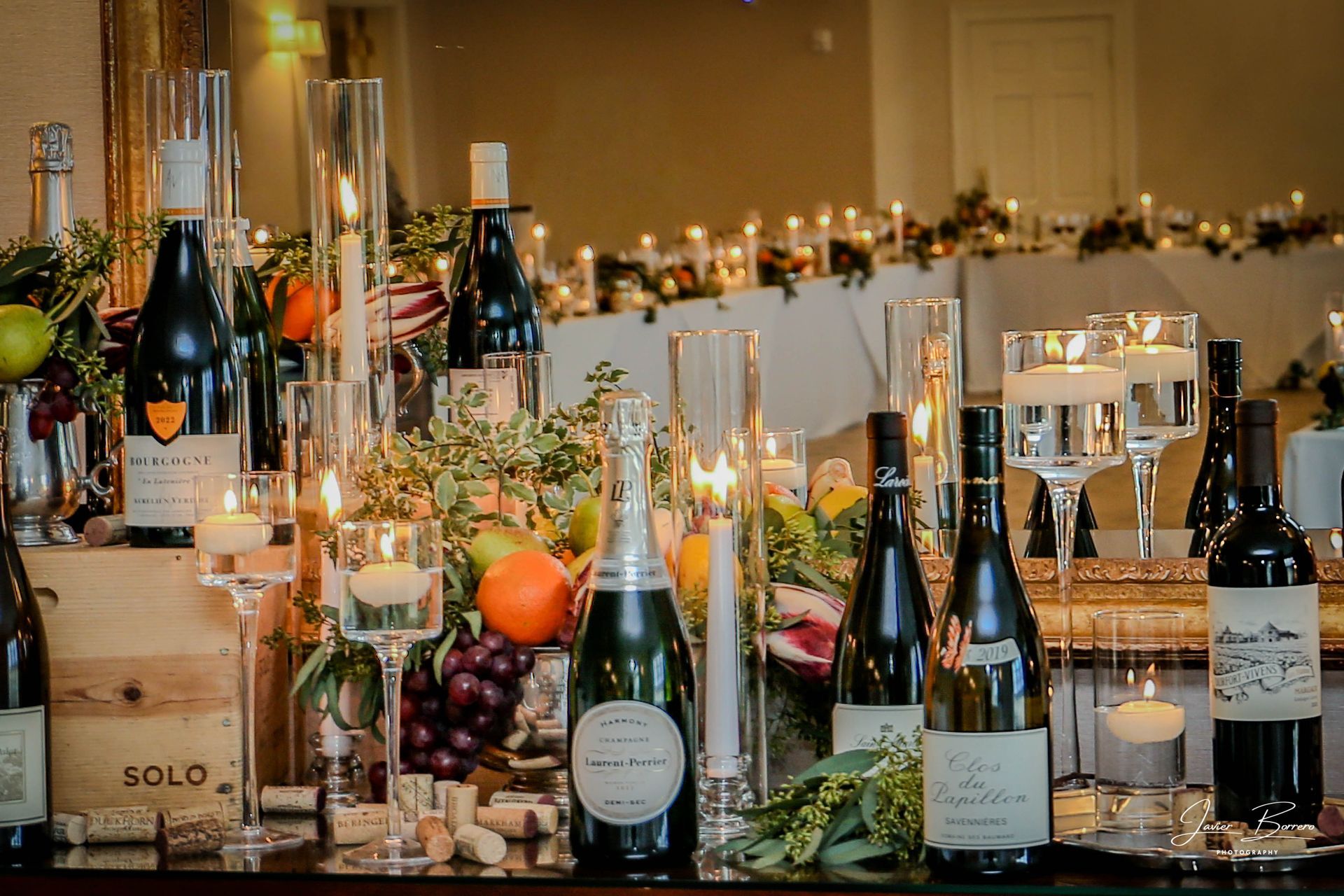 Wine bottles and floating candles on a table decorated with fruits and greenery at an event.