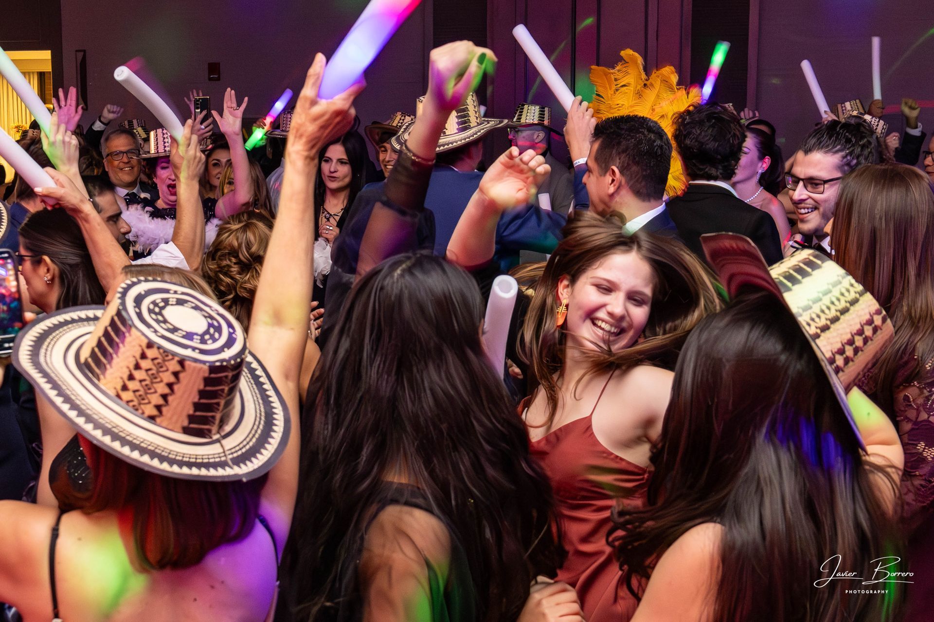 People dance at a celebration, wearing traditional hats and waving glowing foam sticks under colorful party lighting.