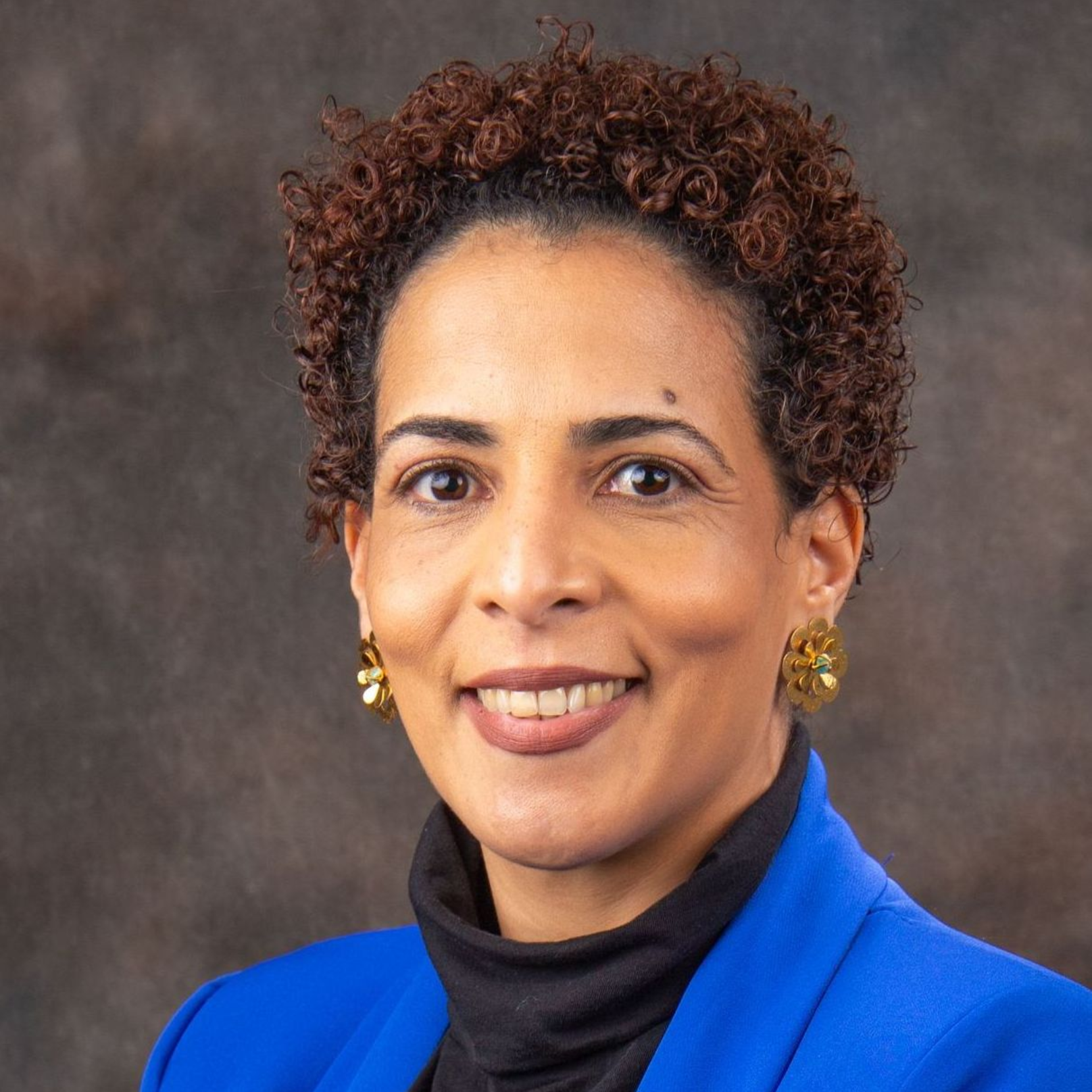 A person with short, curly brown hair wearing a bright blue blazer and a black turtleneck against a gray background.