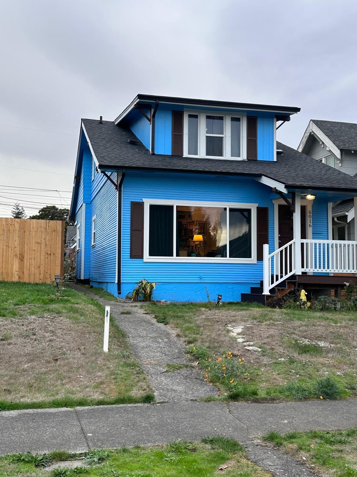 Brown house with white trim and gabled roof, surrounded by lush garden with pink flowers and greenery.