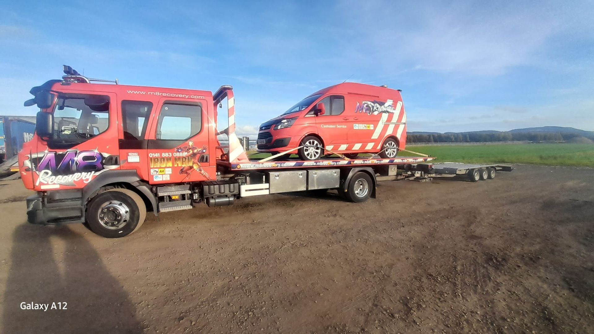 Red tow truck carrying a red van on a flatbed trailer in a field.