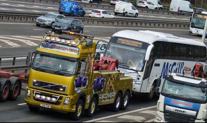 Yellow tow truck towing a white bus on a highway. Other vehicles are in the background.