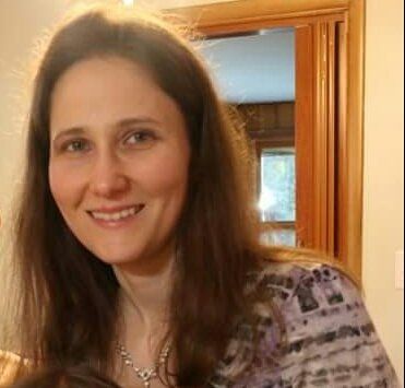 A woman with long brown hair is smiling in front of a door.