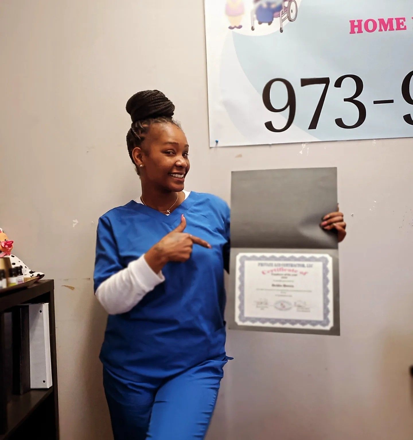 A woman in blue scrubs is holding a certificate and pointing at it