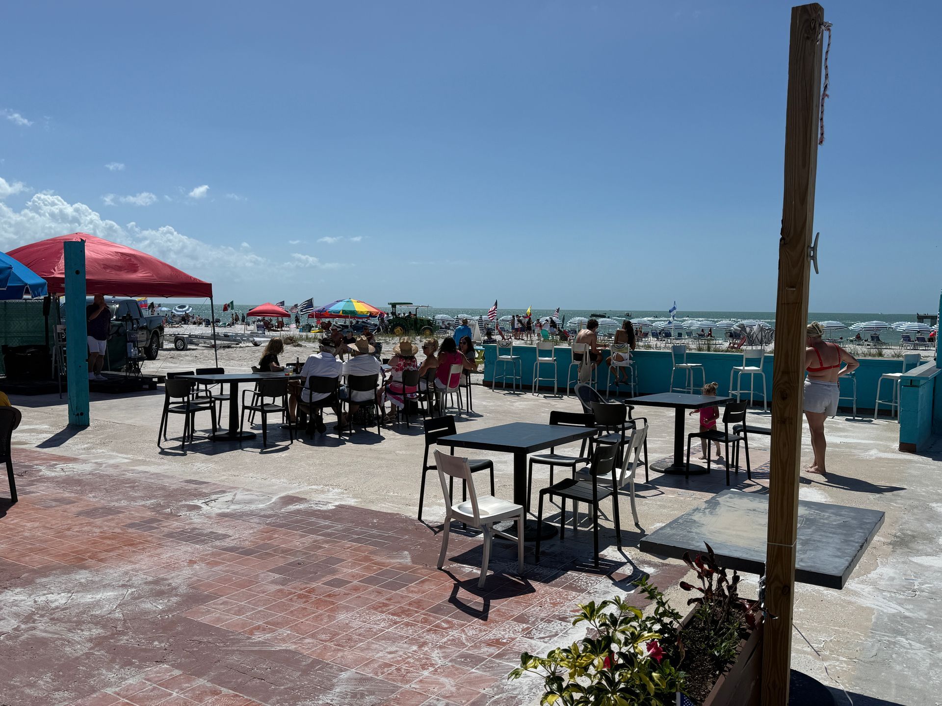 Beachside restaurant with tables, chairs, and a bar, overlooking a crowded sandy beach under a bright blue sky.