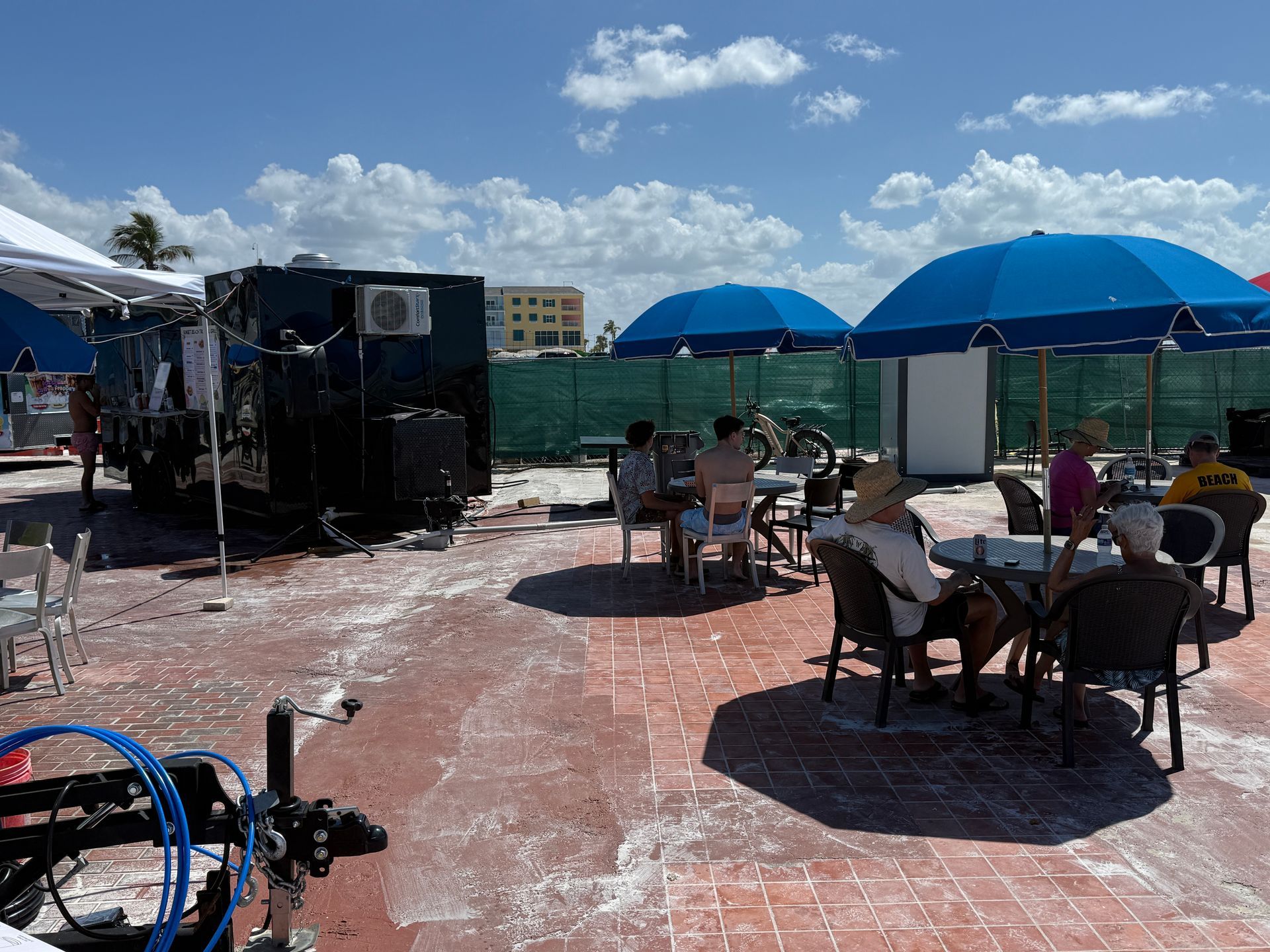 People seated at tables under blue umbrellas, near a food truck and building, on a brick patio.