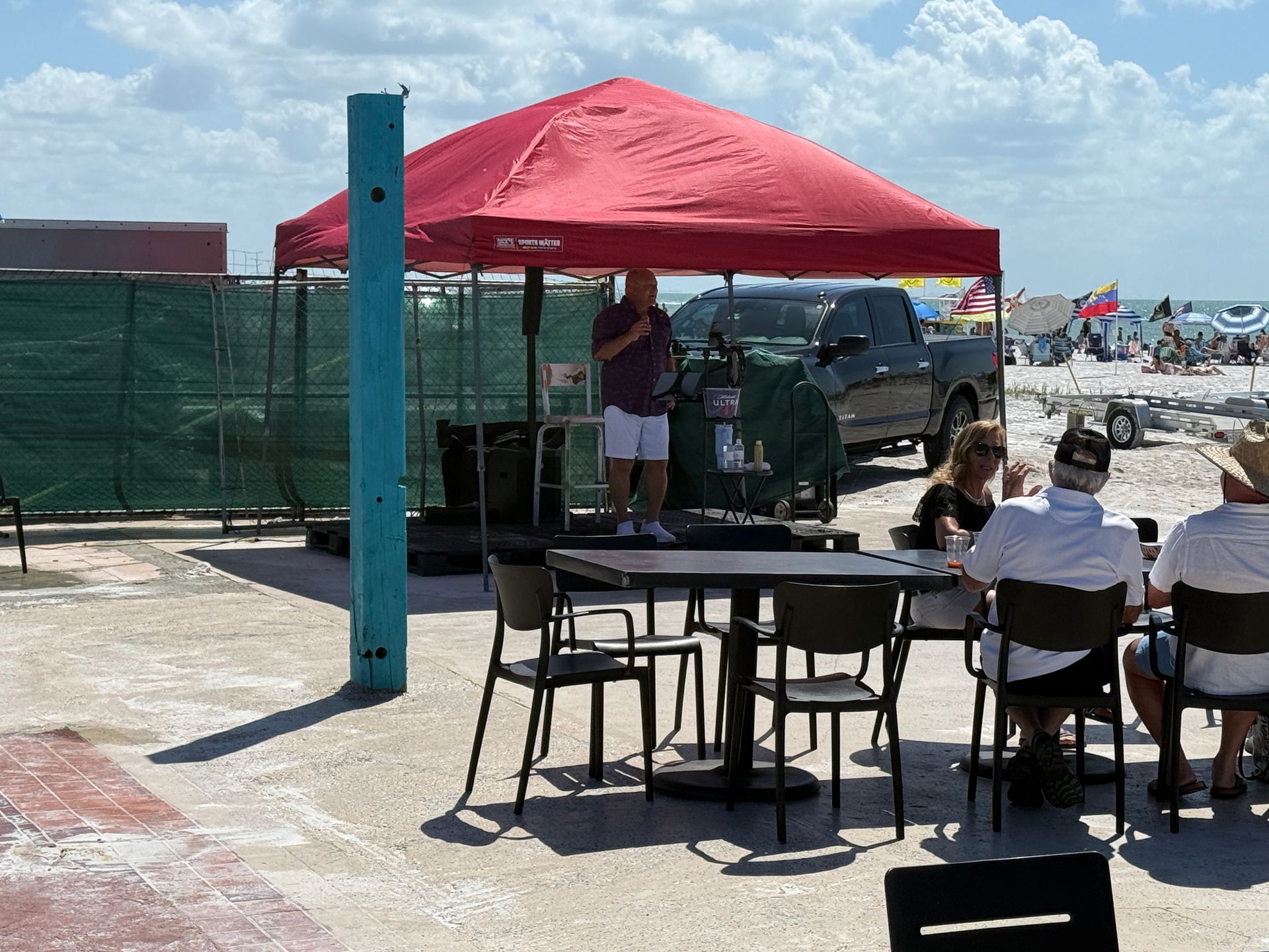 Man speaking under red tent on a beach. People sit at tables nearby. Sunny day.