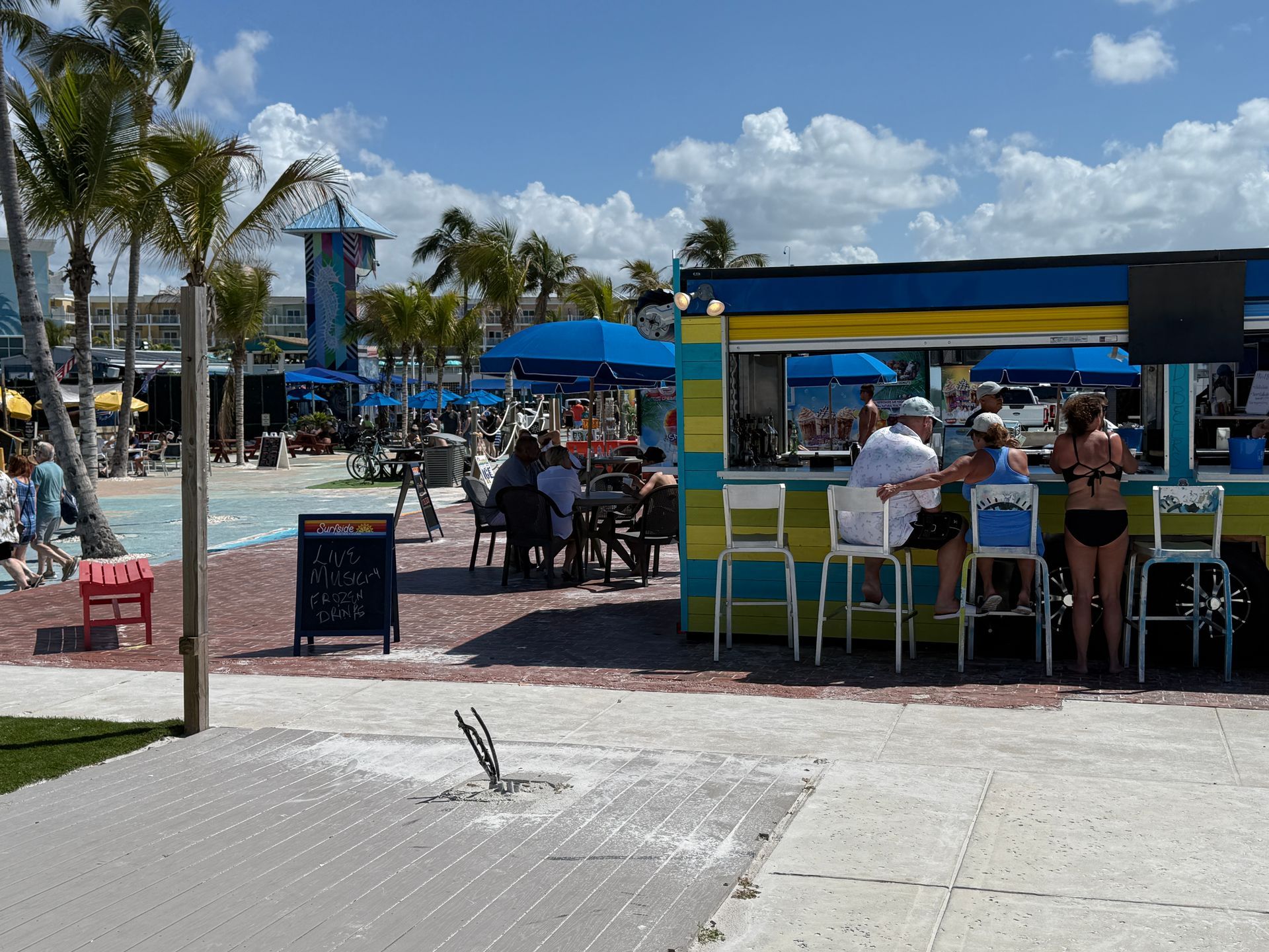 Outdoor bar and seating area. People at tables, bar, and pool. Blue umbrellas, palm trees, sunny day.