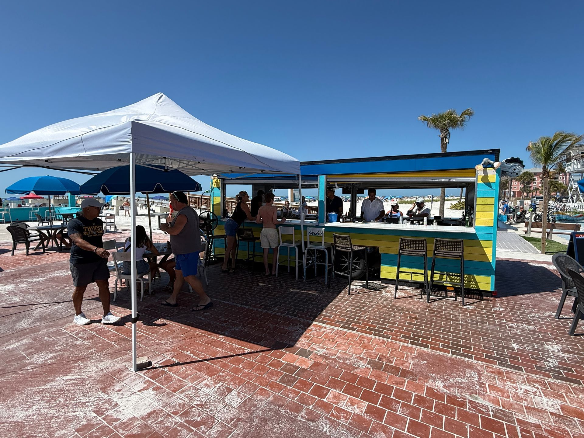 Beachfront bar with people under a white tent and in front of a colorful counter, under a blue sky.