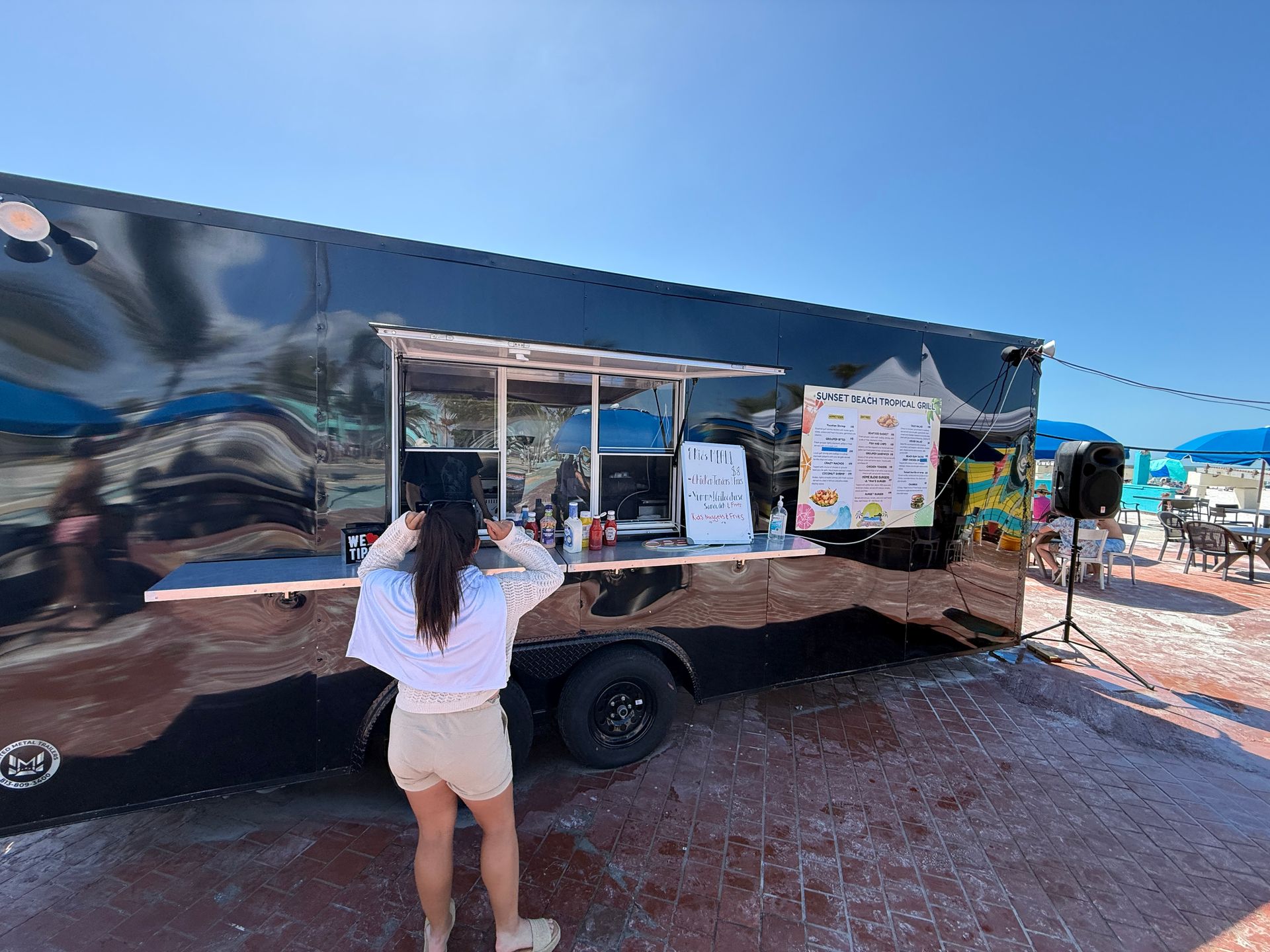 Person in shorts near a black food truck with menu signs on a sunny day.