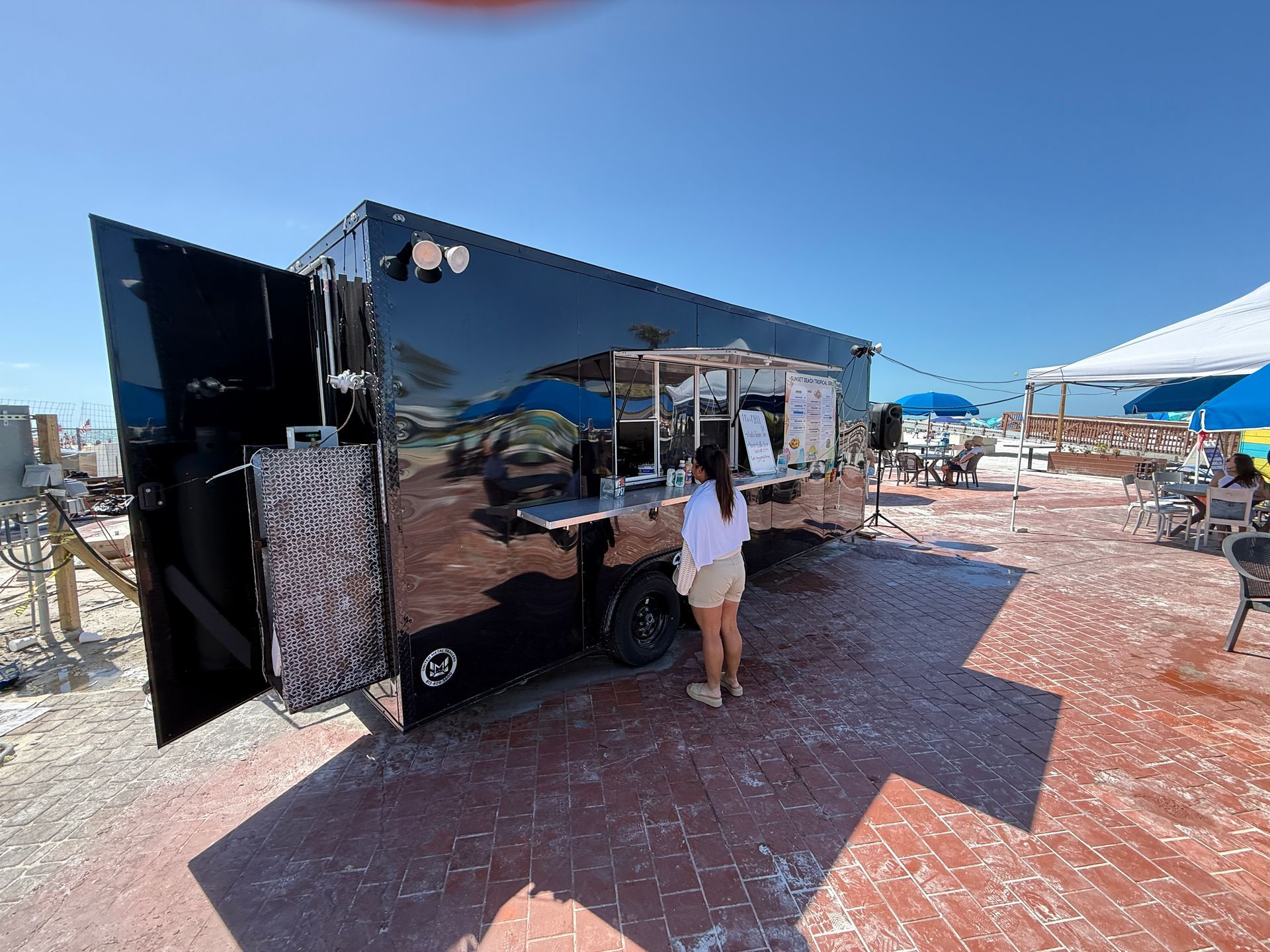 Black food trailer with open door, woman standing at counter, red wood deck, blue sky.