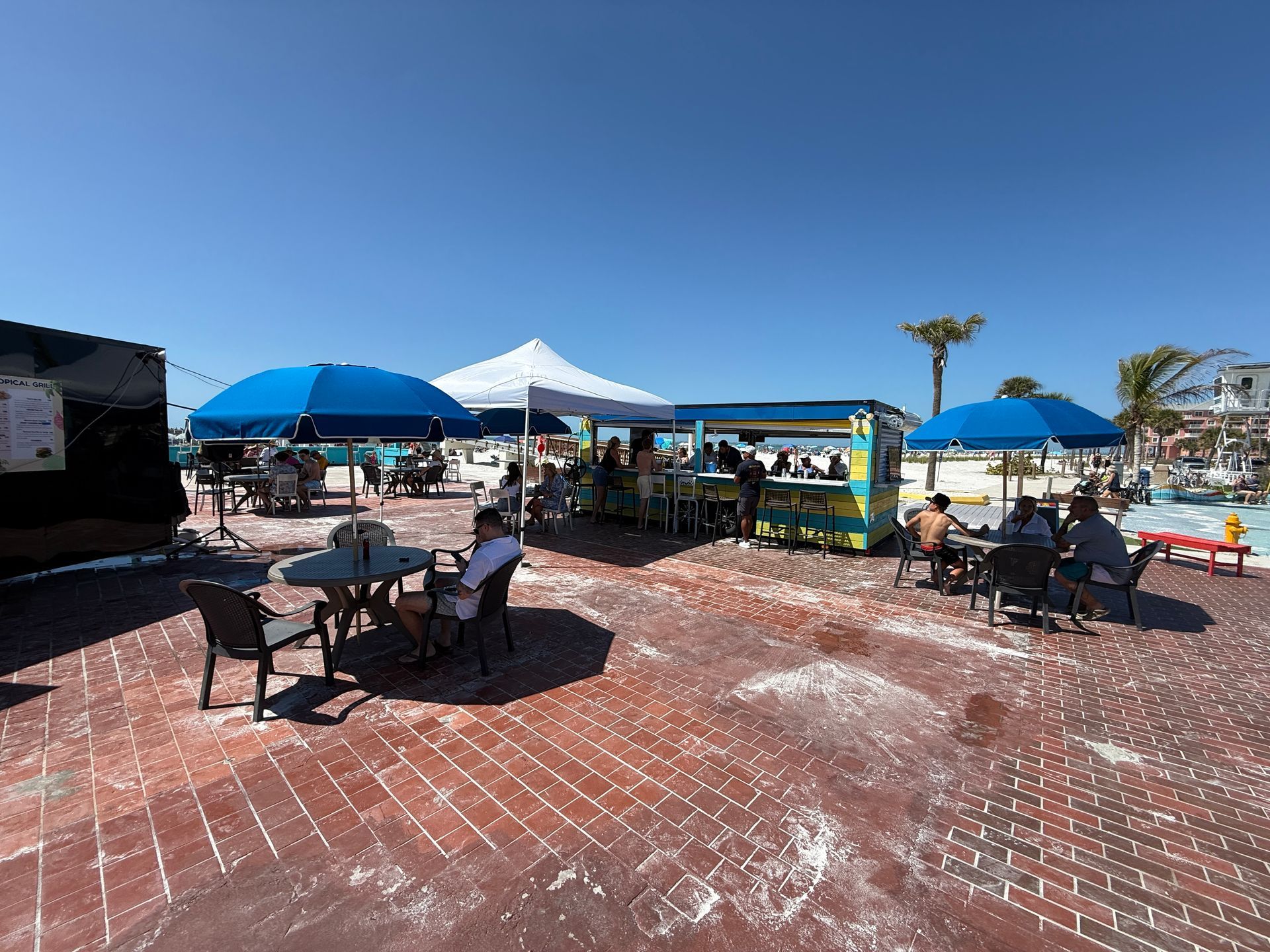 Outdoor bar with tables, umbrellas, and people under a bright blue sky.