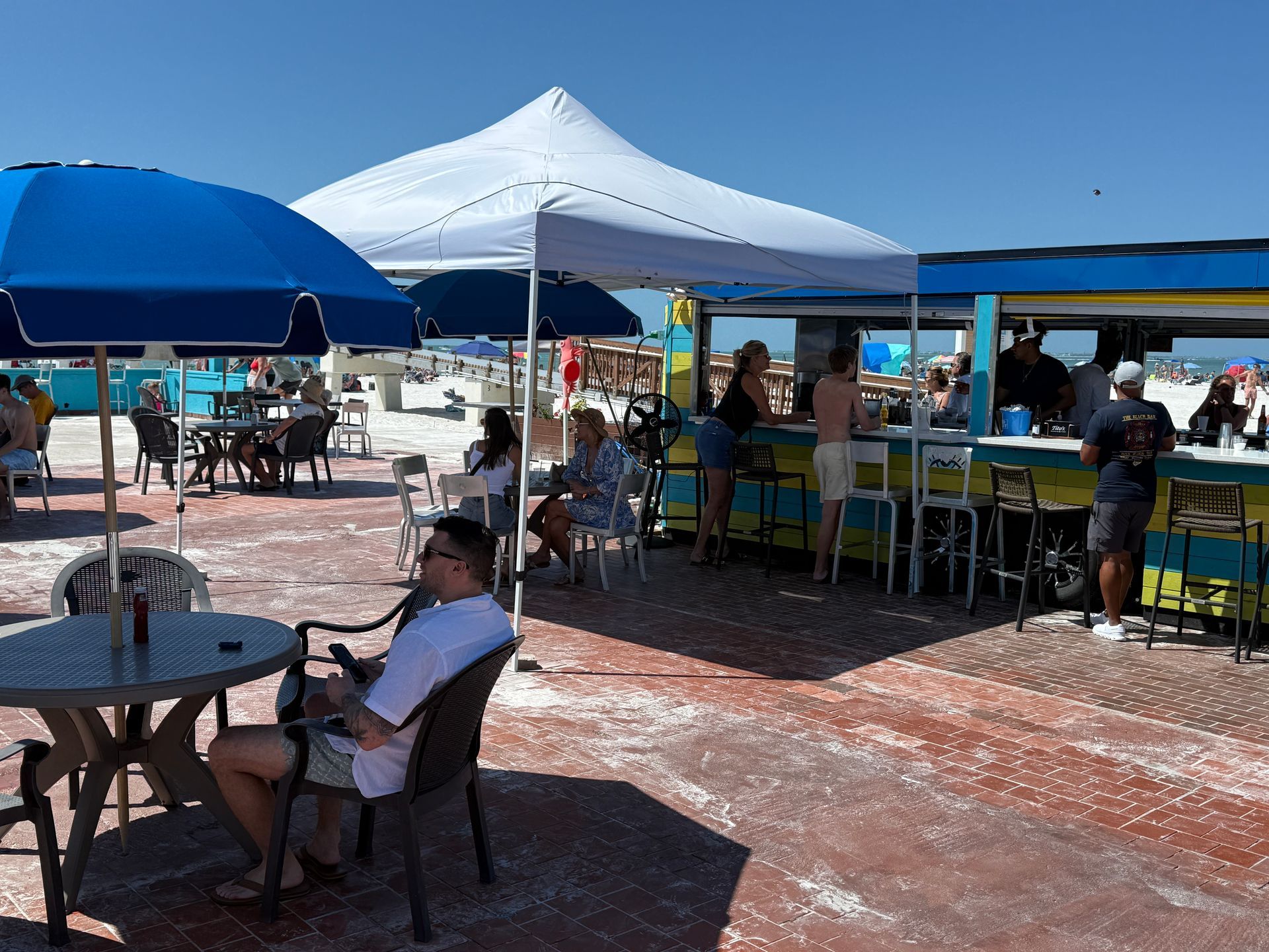 Outdoor beach bar with patrons seated at tables and bar, beneath umbrellas and canopies on a sunny day.