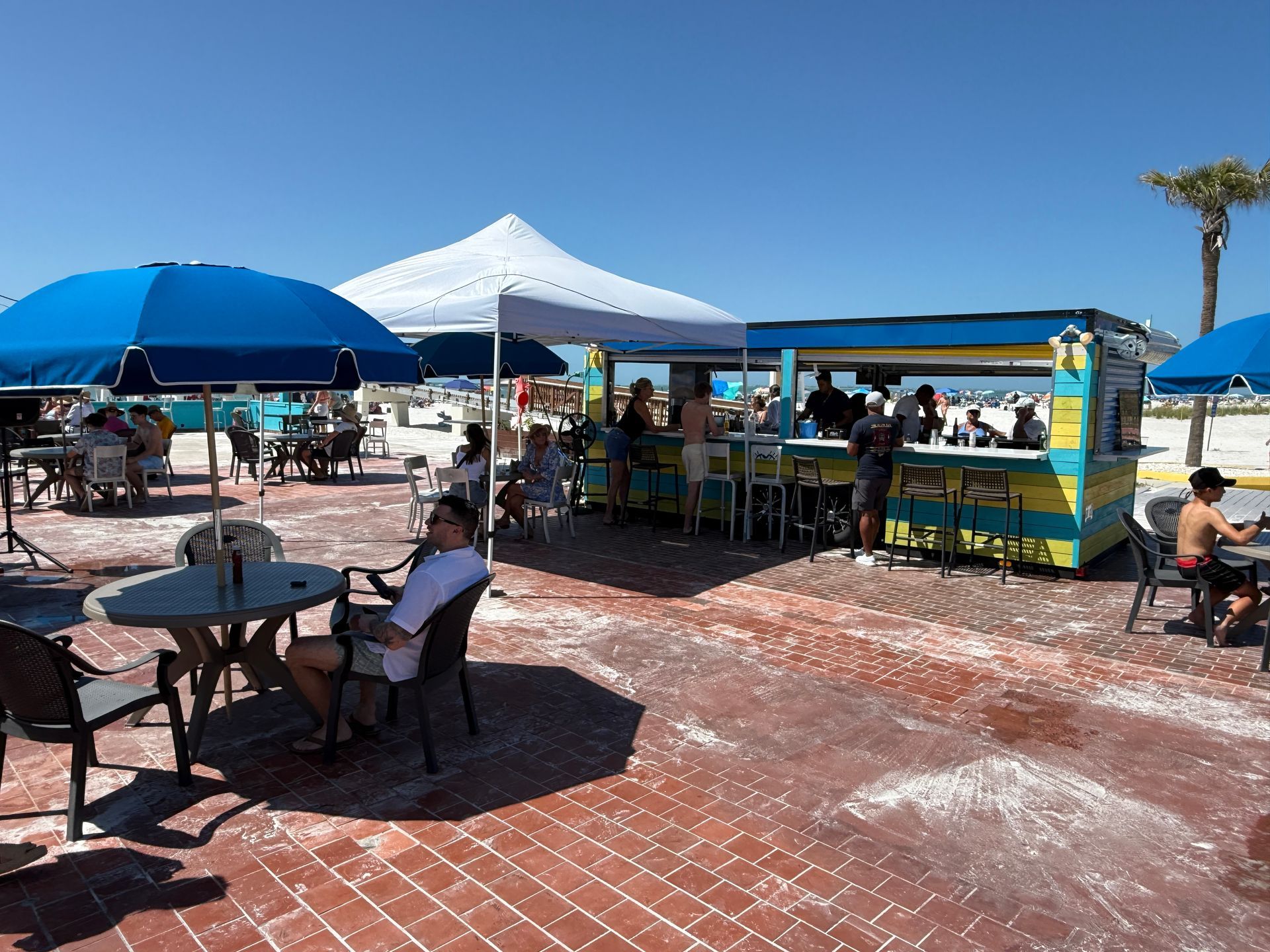Beachside bar with patrons under umbrellas, a tent, and a brightly colored bar structure.
