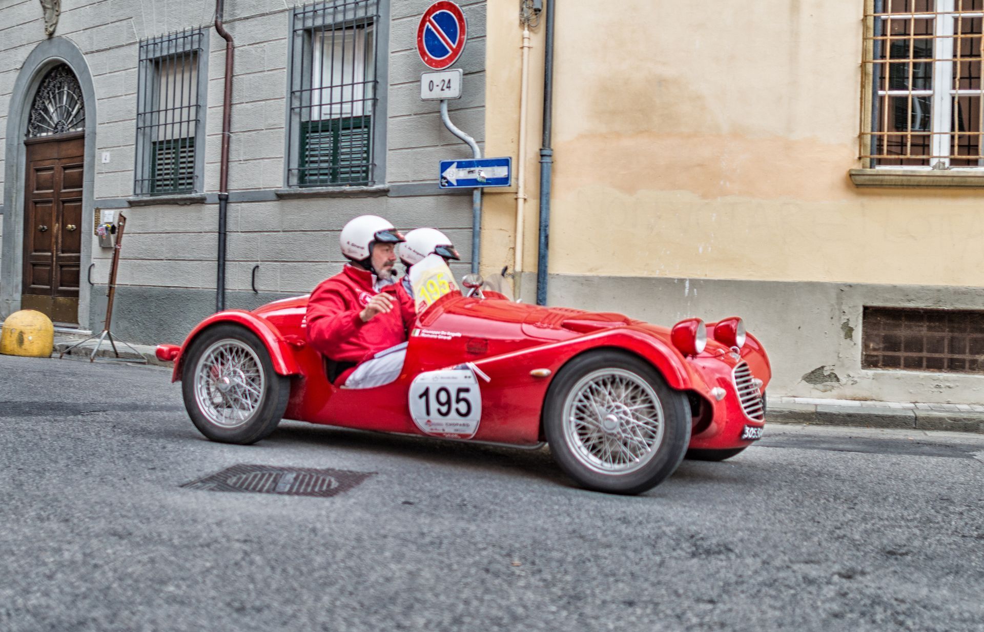 Auto da corsa rossa d'epoca con due persone che indossano il casco che guidano su una strada cittadina; numero 195 sull'auto.
