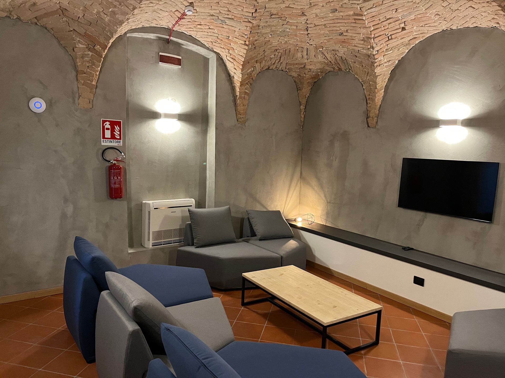 Accogliente zona lounge con divani grigi, TV e soffitto in mattoni, dotata di estintore e rilevatore di fumo.