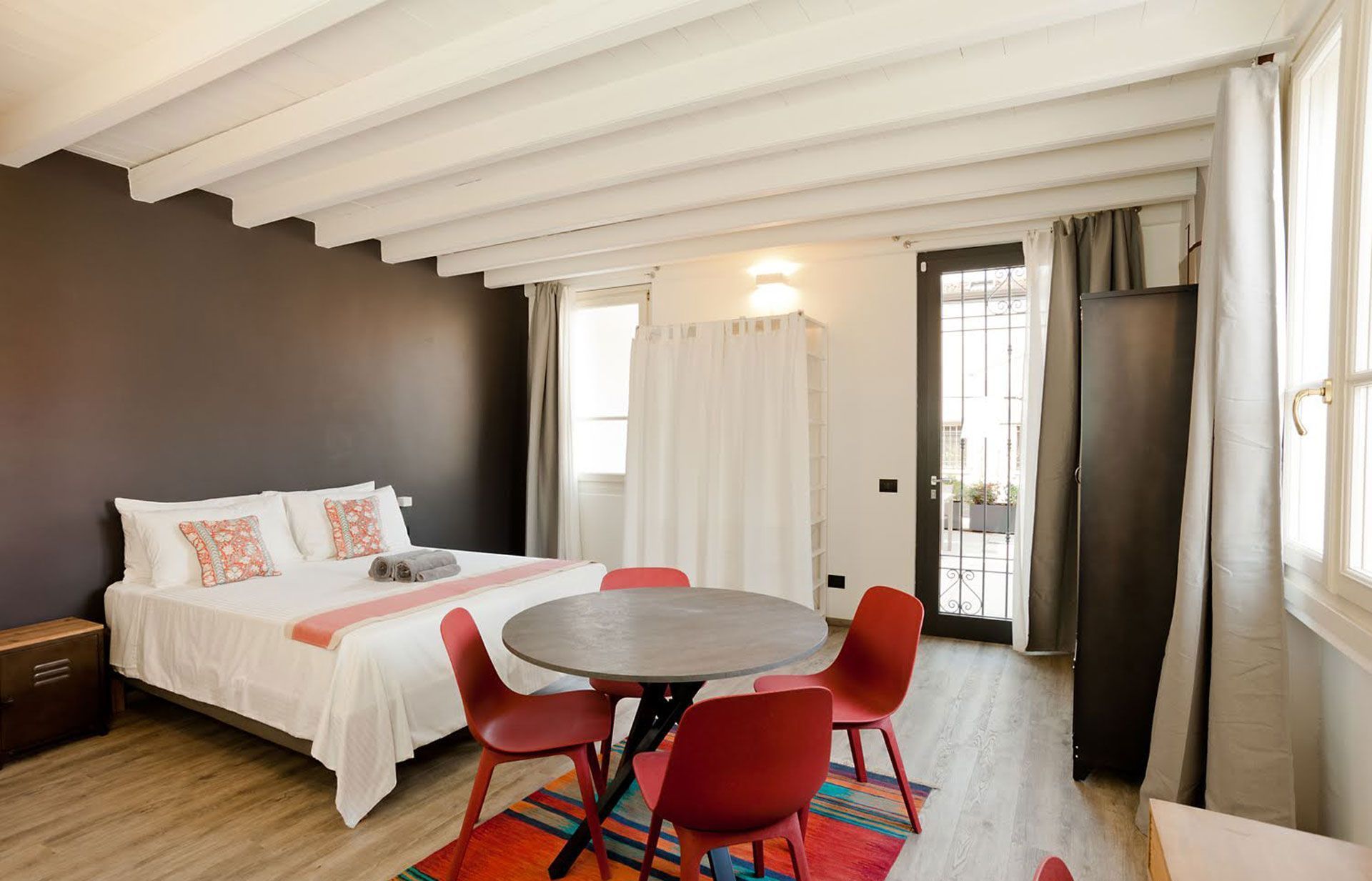 Camera da letto con letto matrimoniale, tavolo rotondo con sedie rosse, tappeto, parete scura e travi in ​​legno.