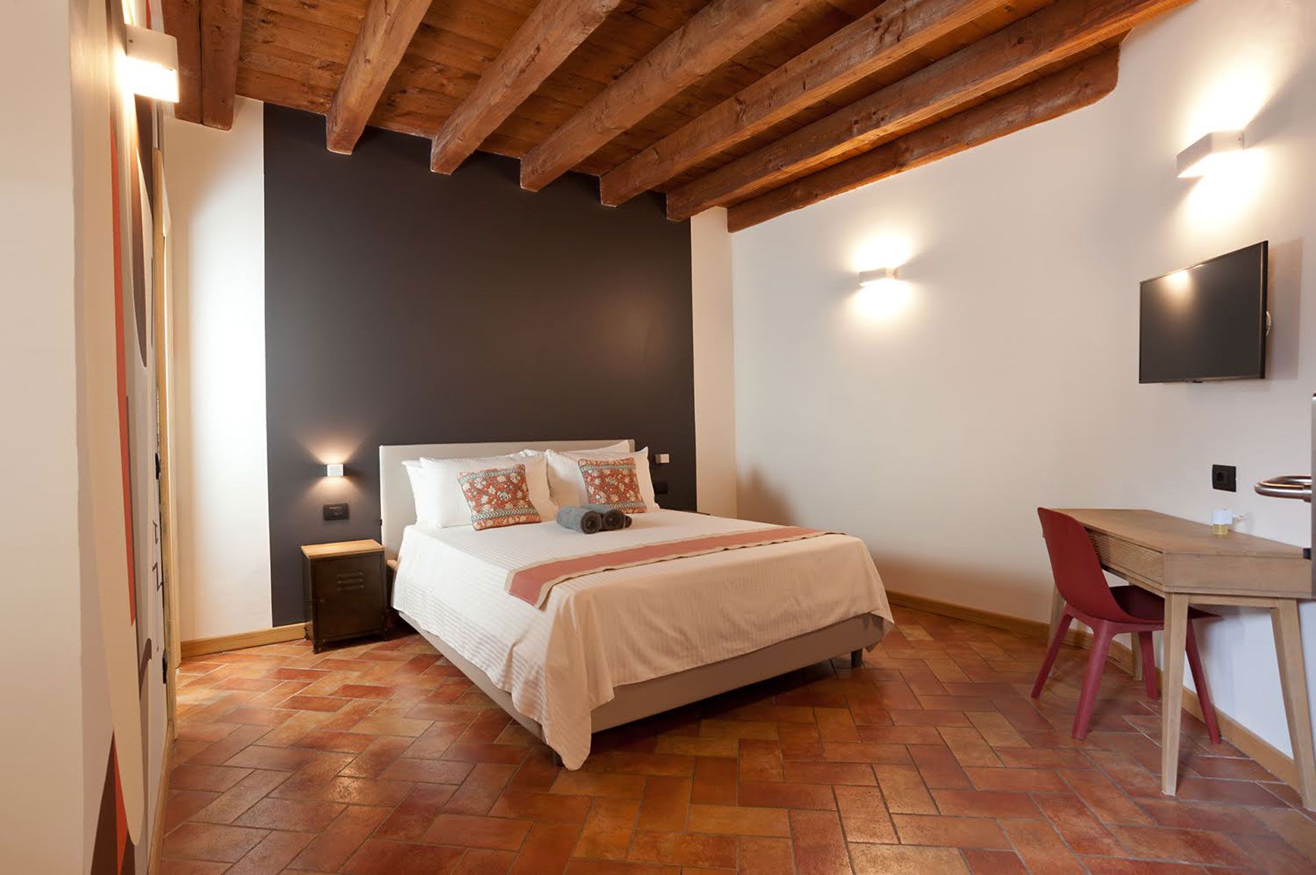 Camera da letto con letto, scrivania e soffitto in legno a vista. Parete decorativa grigio scuro, pavimento in cotto.