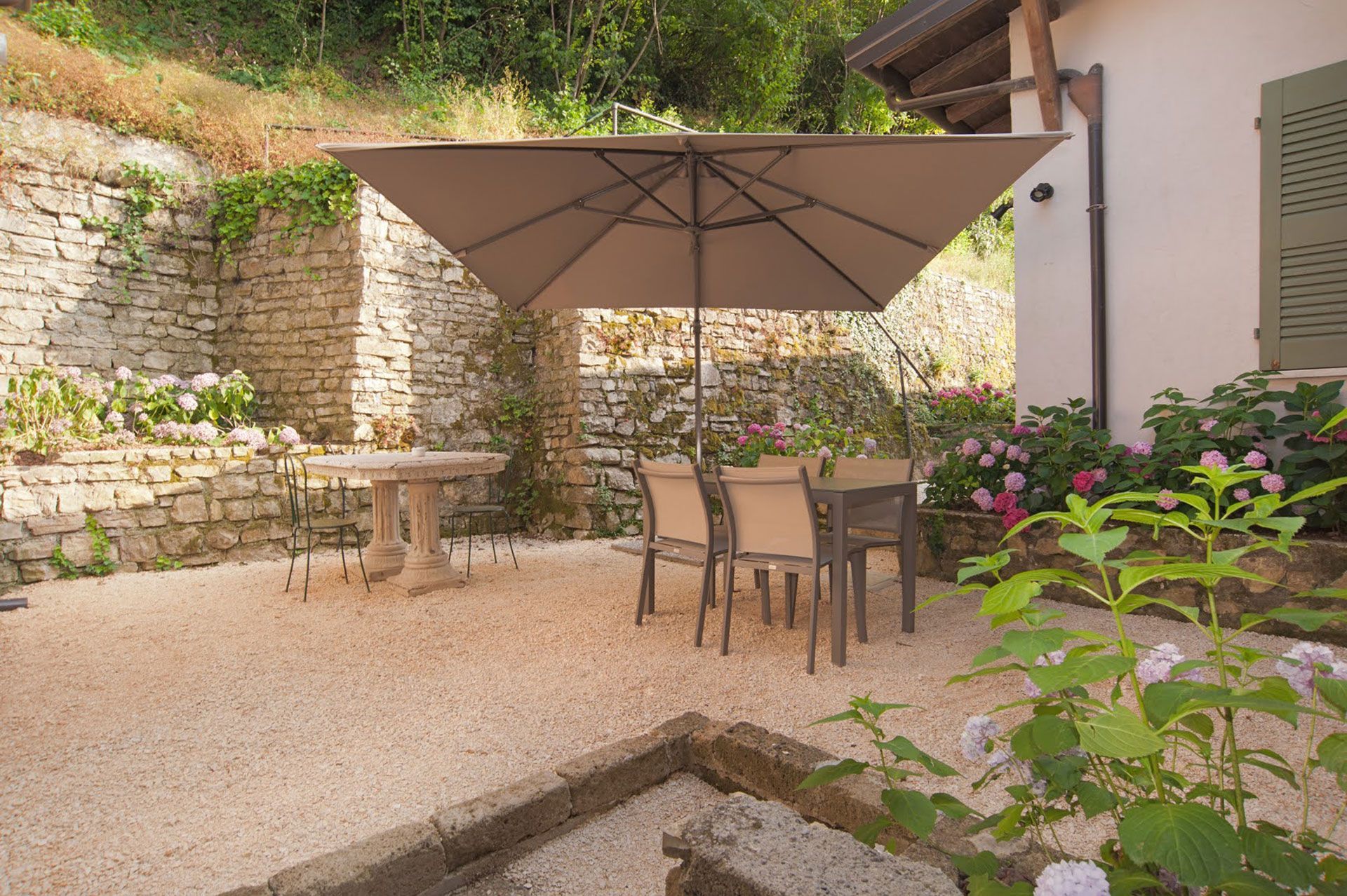 Patio in pietra con zona pranzo all'aperto sotto un grande ombrellone color marrone chiaro, con muri in pietra e fiori.