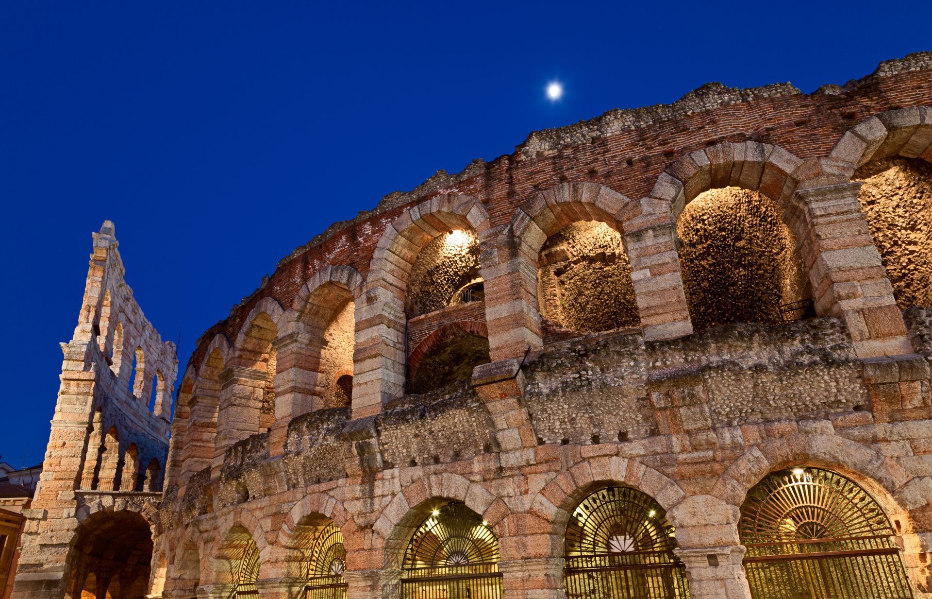 L'Arena di Verona illuminata di notte sotto un cielo blu scuro con una luna crescente.