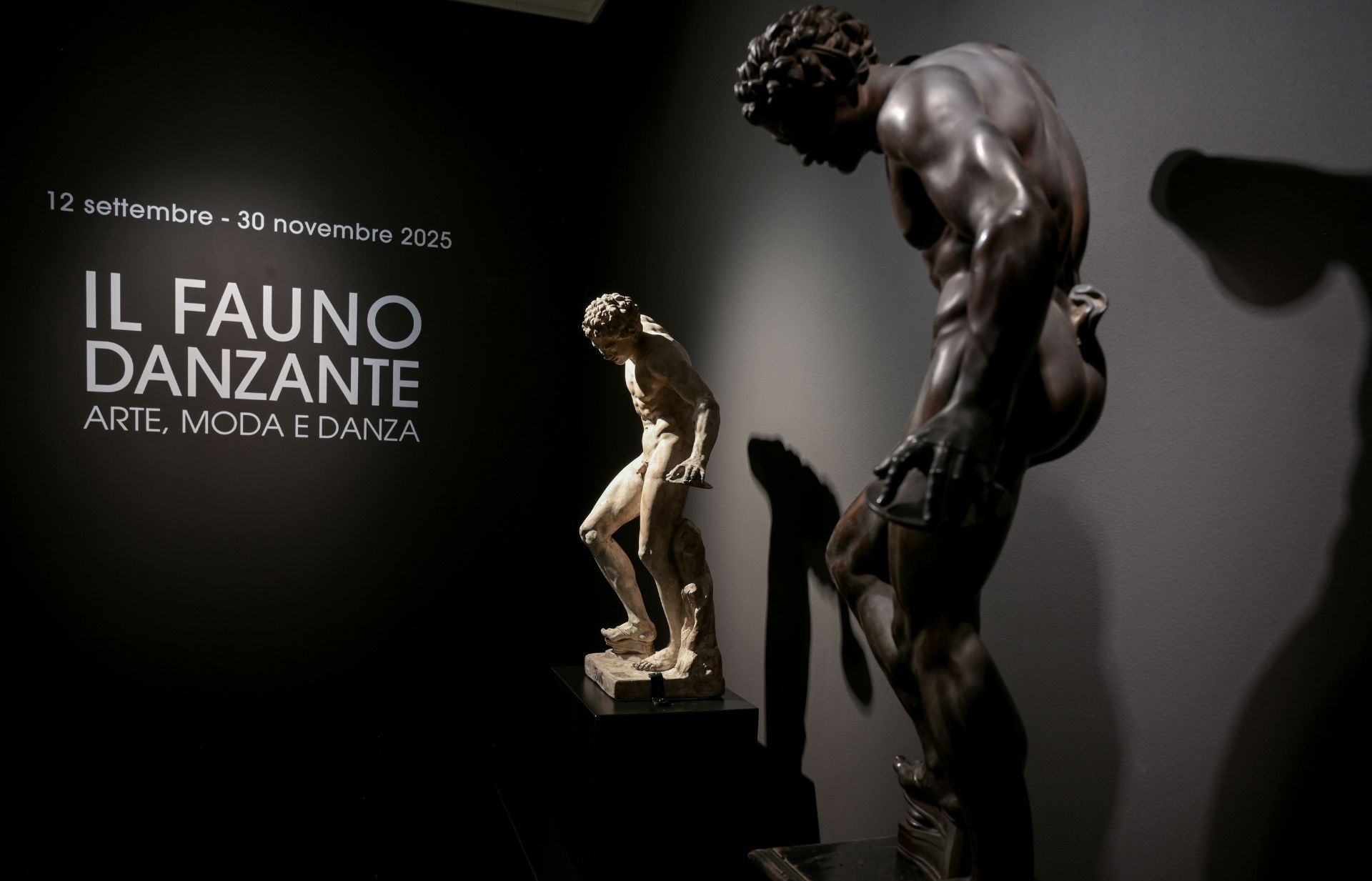 Two bronze statues of male figures in a dark gallery; one is large and bent over, the other is smaller and standing.