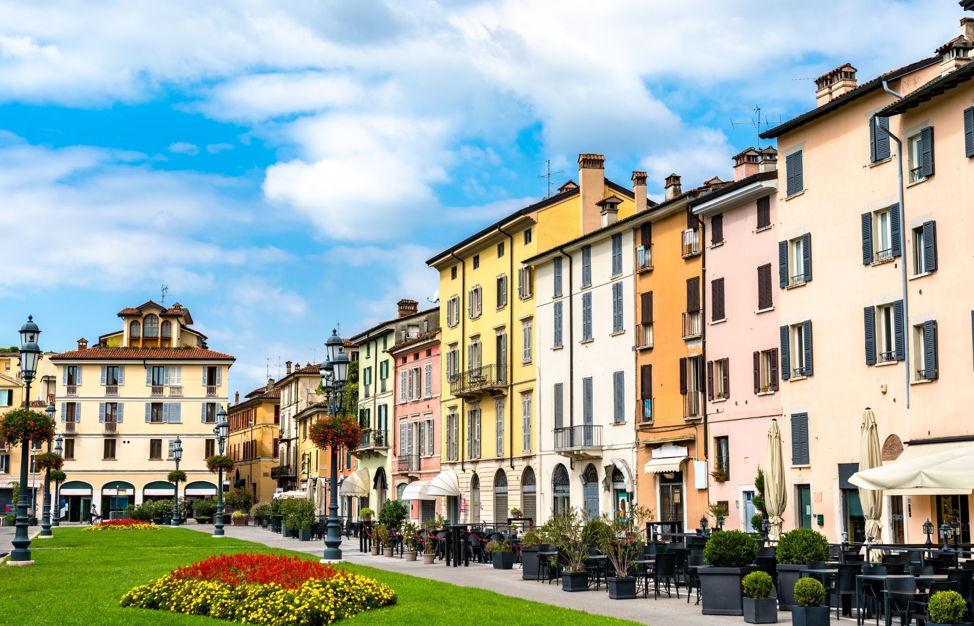 Una vivace piazza di una cittadina europea con edifici colorati, un prato ben curato, aiuole fiorite e tavolini all'aperto per i clienti del caffè.