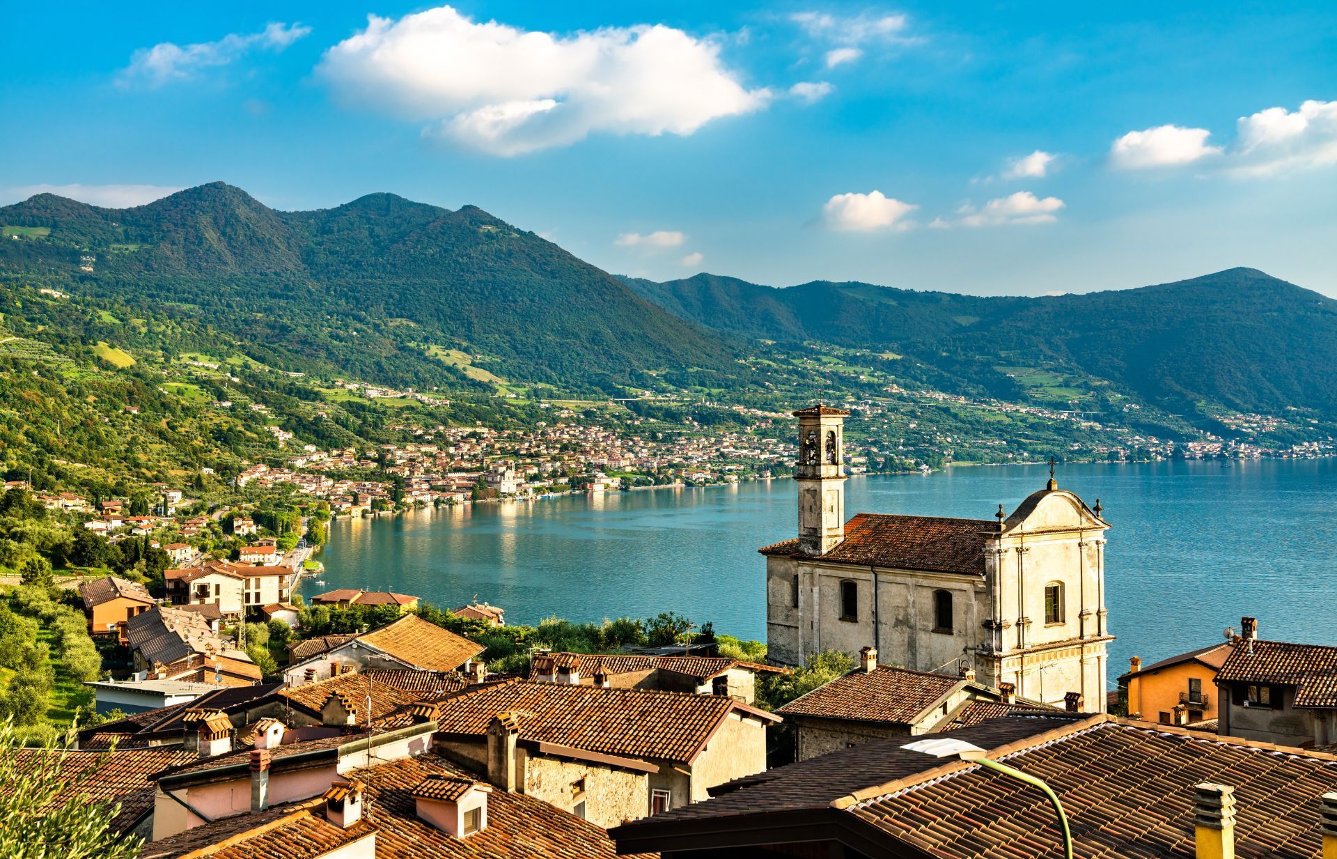 Una storica chiesa in pietra si affaccia su un tranquillo lago azzurro, circondato da lussureggianti montagne verdi, sotto un cielo luminoso e nuvoloso.