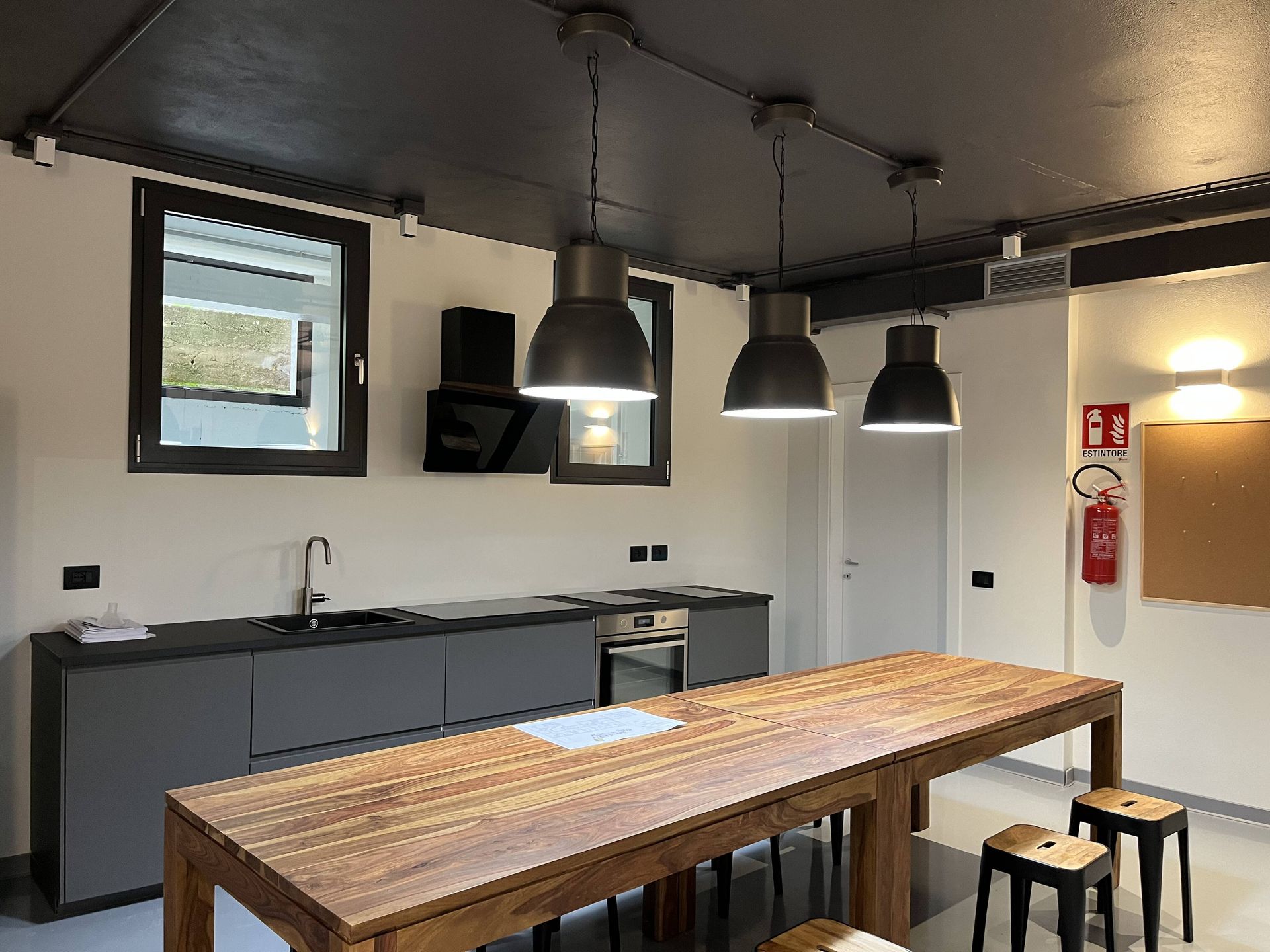 Cucina con isola e mobili grigi, tre lampade a sospensione. Finestre, elettrodomestici e sgabelli presenti.